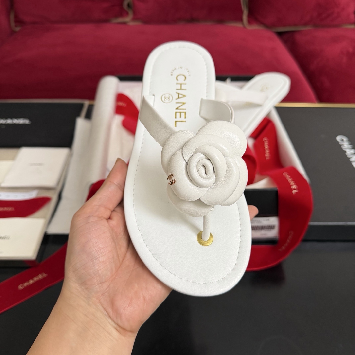A pair of luxury white Chanel thong sandals featuring a large camellia flower with a gold CC logo on the strap, presented in a black box with red ribbons. - Hình 3