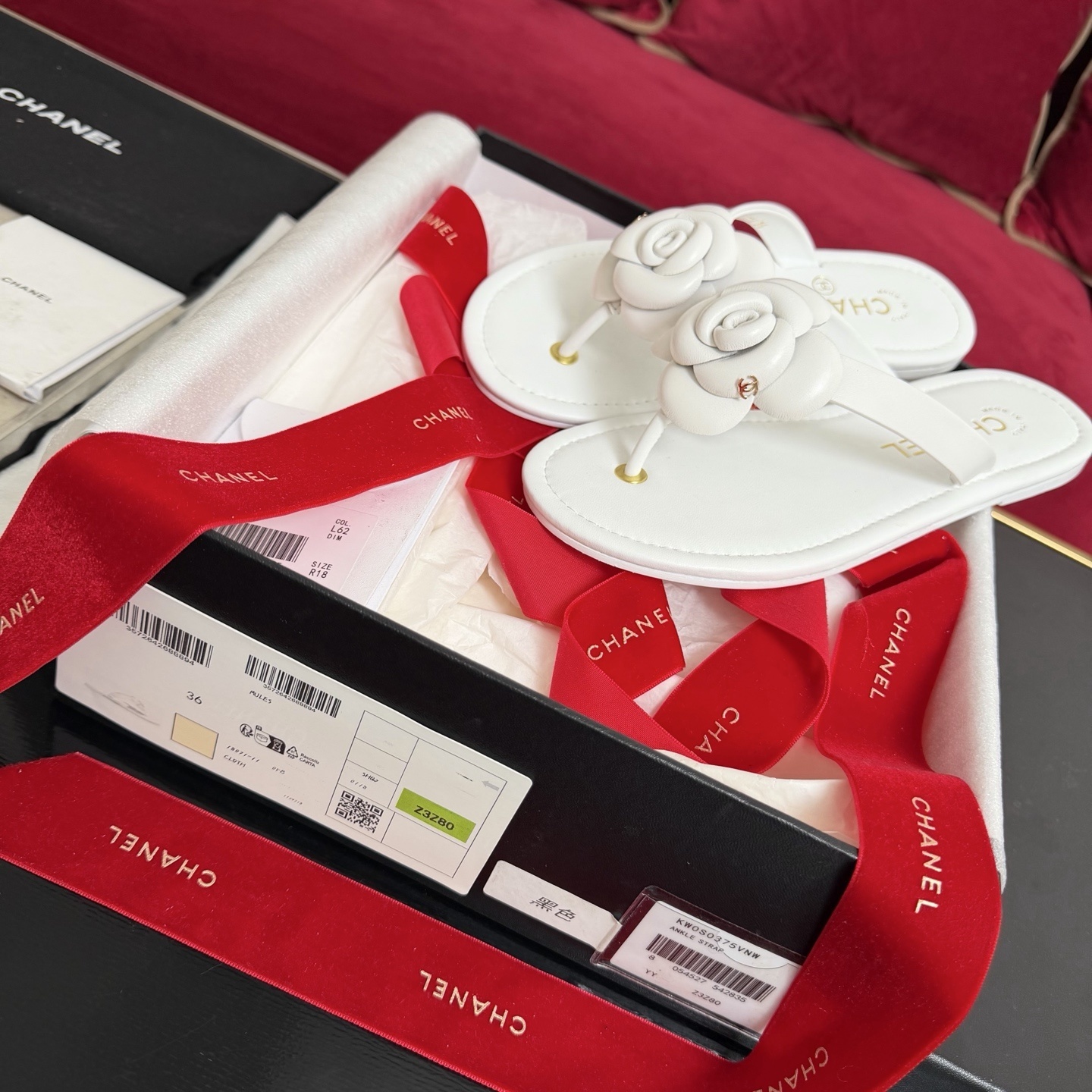 A pair of luxury white Chanel thong sandals featuring a large camellia flower with a gold CC logo on the strap, presented in a black box with red ribbons. - Hình 2
