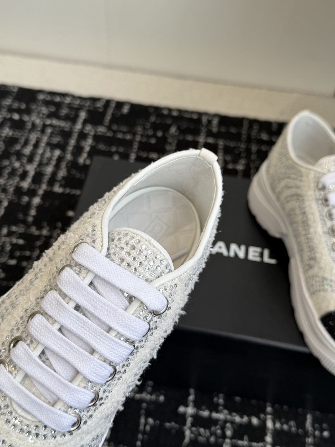 A pair of luxury white tweed sneakers with black rhinestone cap toes and chunky white soles, presented on a black designer shoe box. - Hình 7