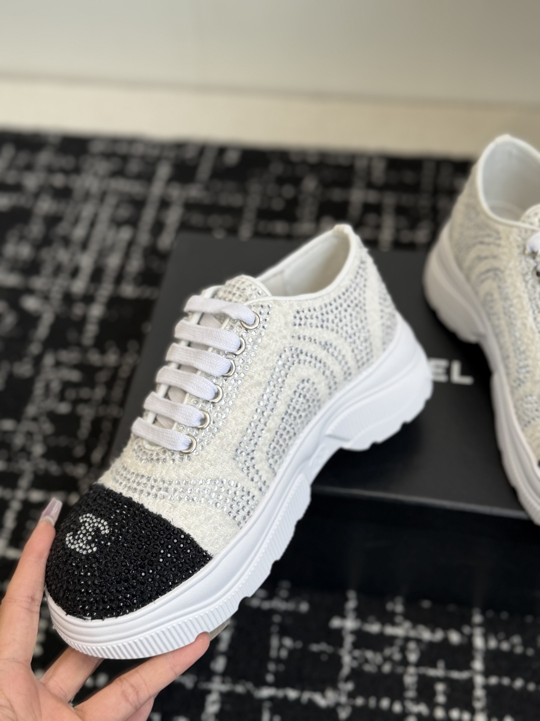 A pair of luxury white tweed sneakers with black rhinestone cap toes and chunky white soles, presented on a black designer shoe box. - Hình 6