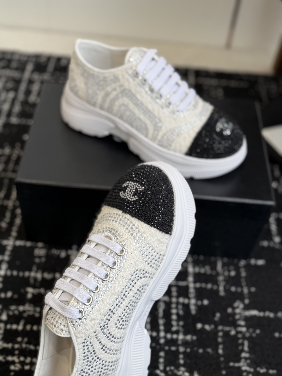 A pair of luxury white tweed sneakers with black rhinestone cap toes and chunky white soles, presented on a black designer shoe box. - Hình 5