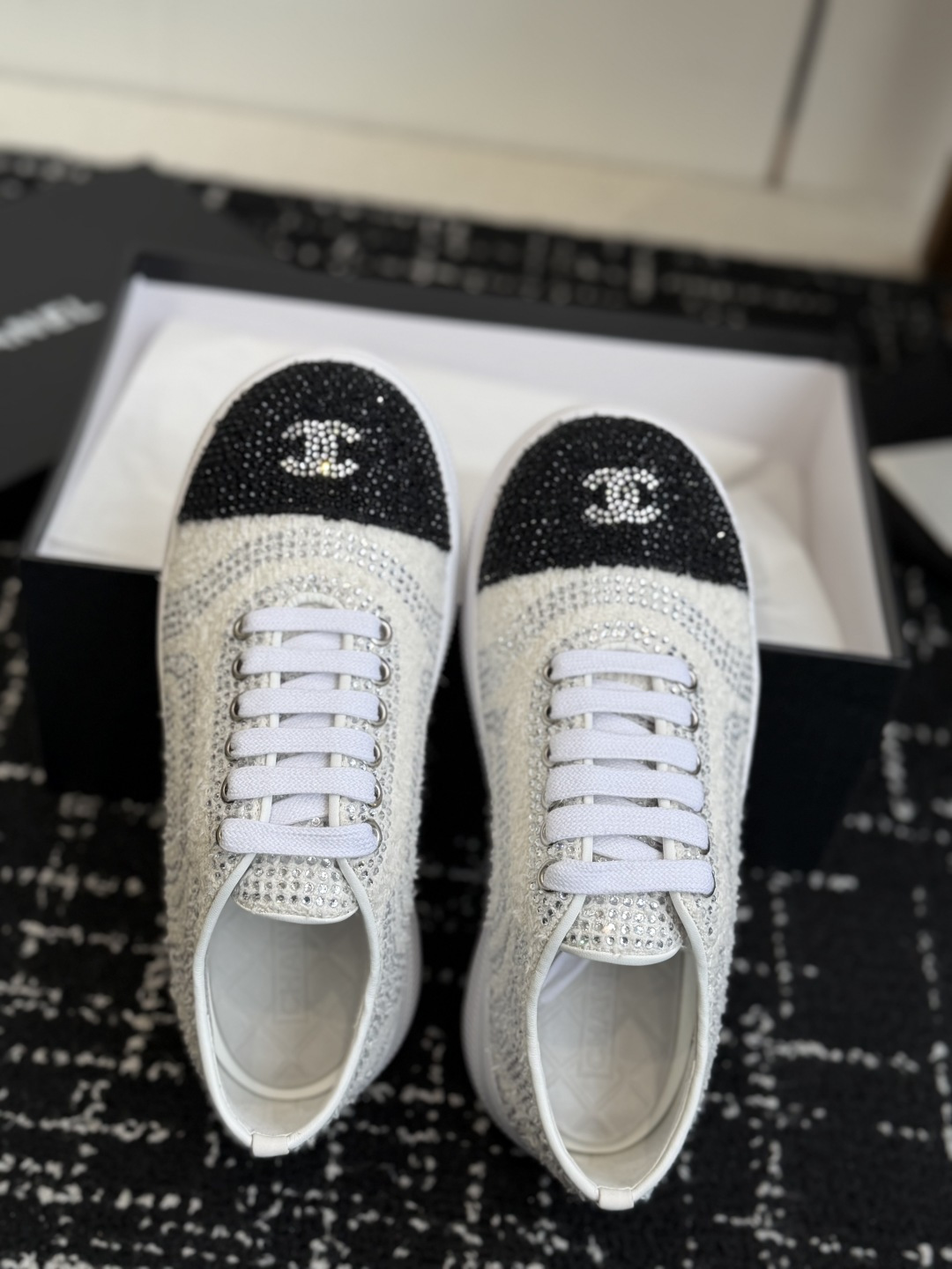 A pair of luxury white tweed sneakers with black rhinestone cap toes and chunky white soles, presented on a black designer shoe box. - Hình 4