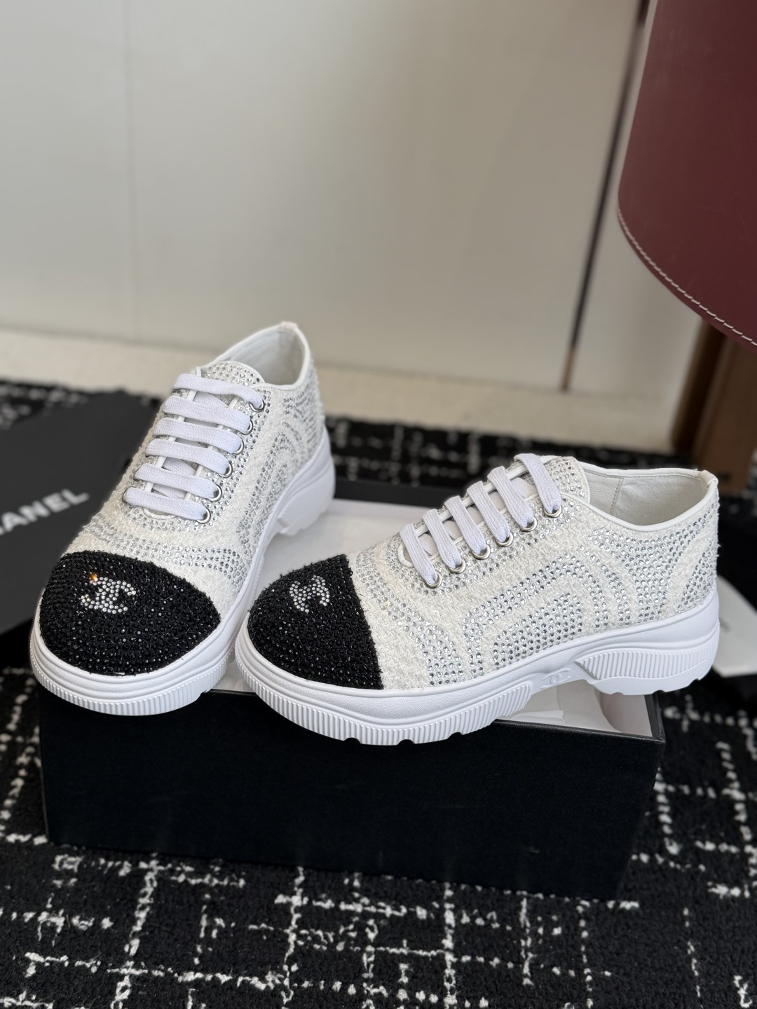 A pair of luxury white tweed sneakers with black rhinestone cap toes and chunky white soles, presented on a black designer shoe box. - Hình 3