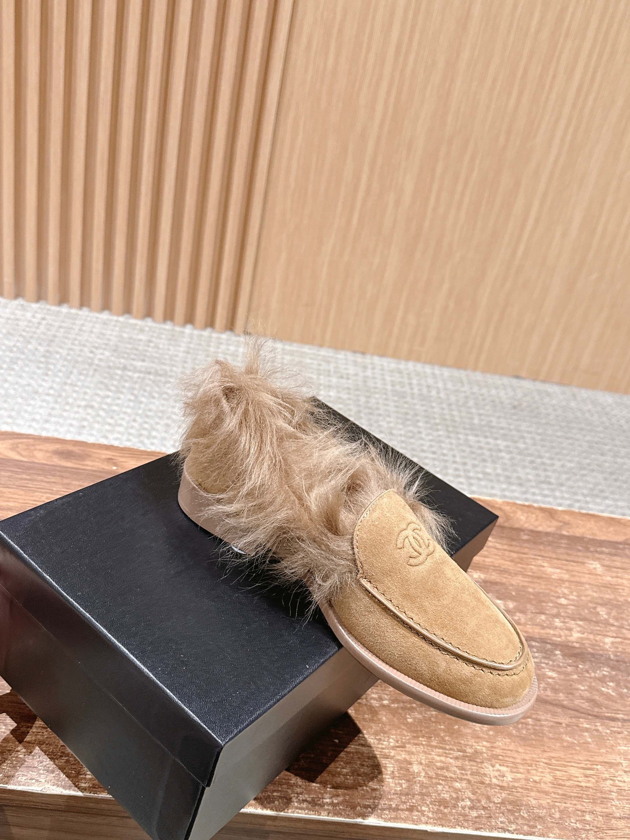 A pair of tan Chanel suede loafers with fluffy brown fur trim and embossed CC logo on a wooden surface next to a black shoe box. - Hình 3