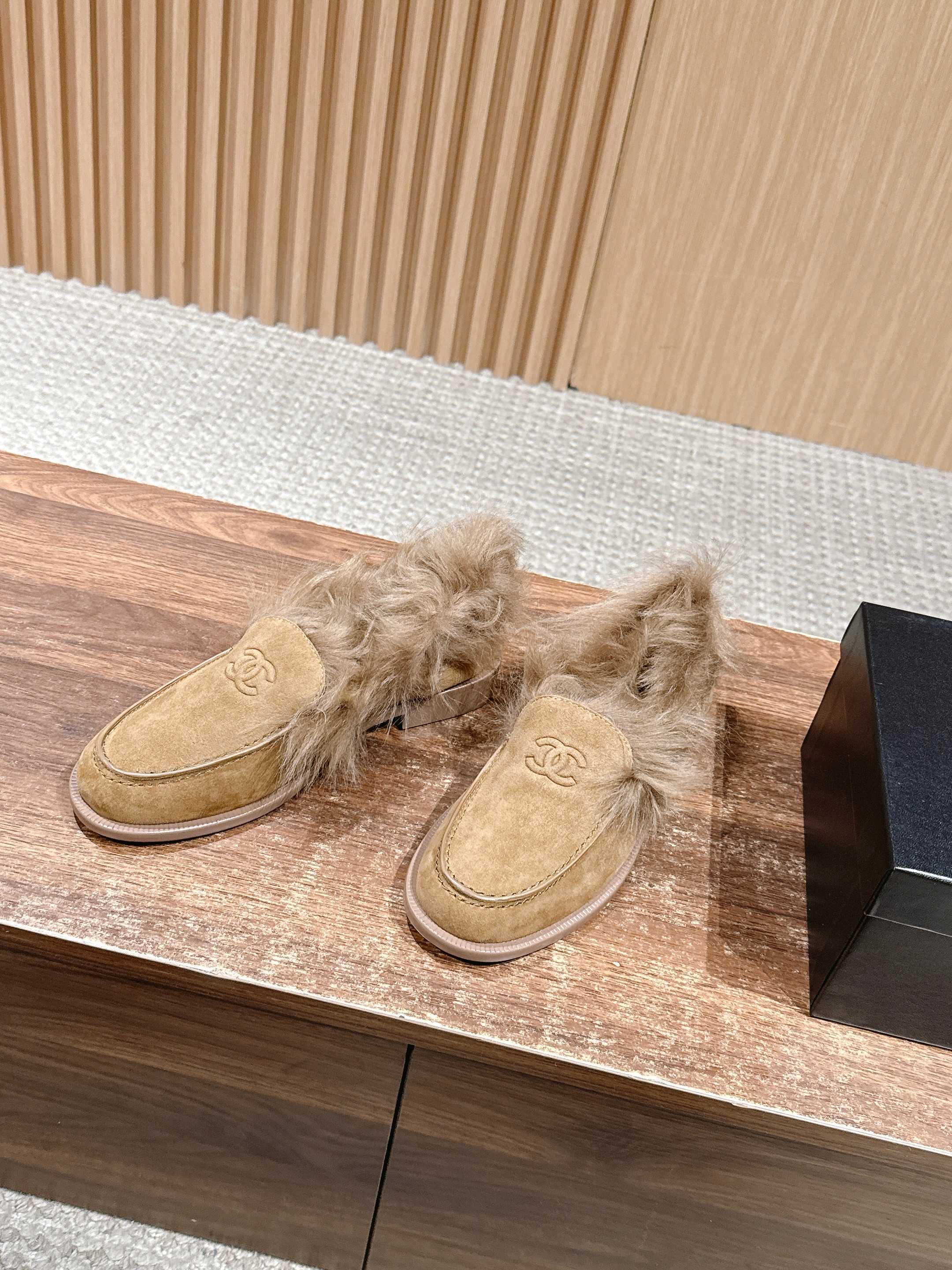 A pair of tan Chanel suede loafers with fluffy brown fur trim and embossed CC logo on a wooden surface next to a black shoe box. - Hình 2