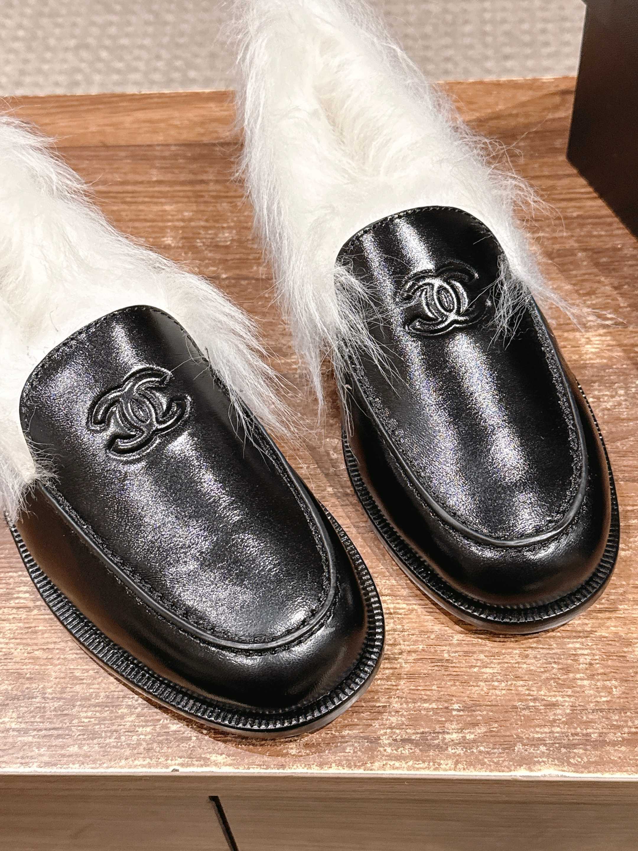 A pair of luxury black leather Chanel loafers with prominent white fur lining and CC logo on a wooden surface. - Hình 6