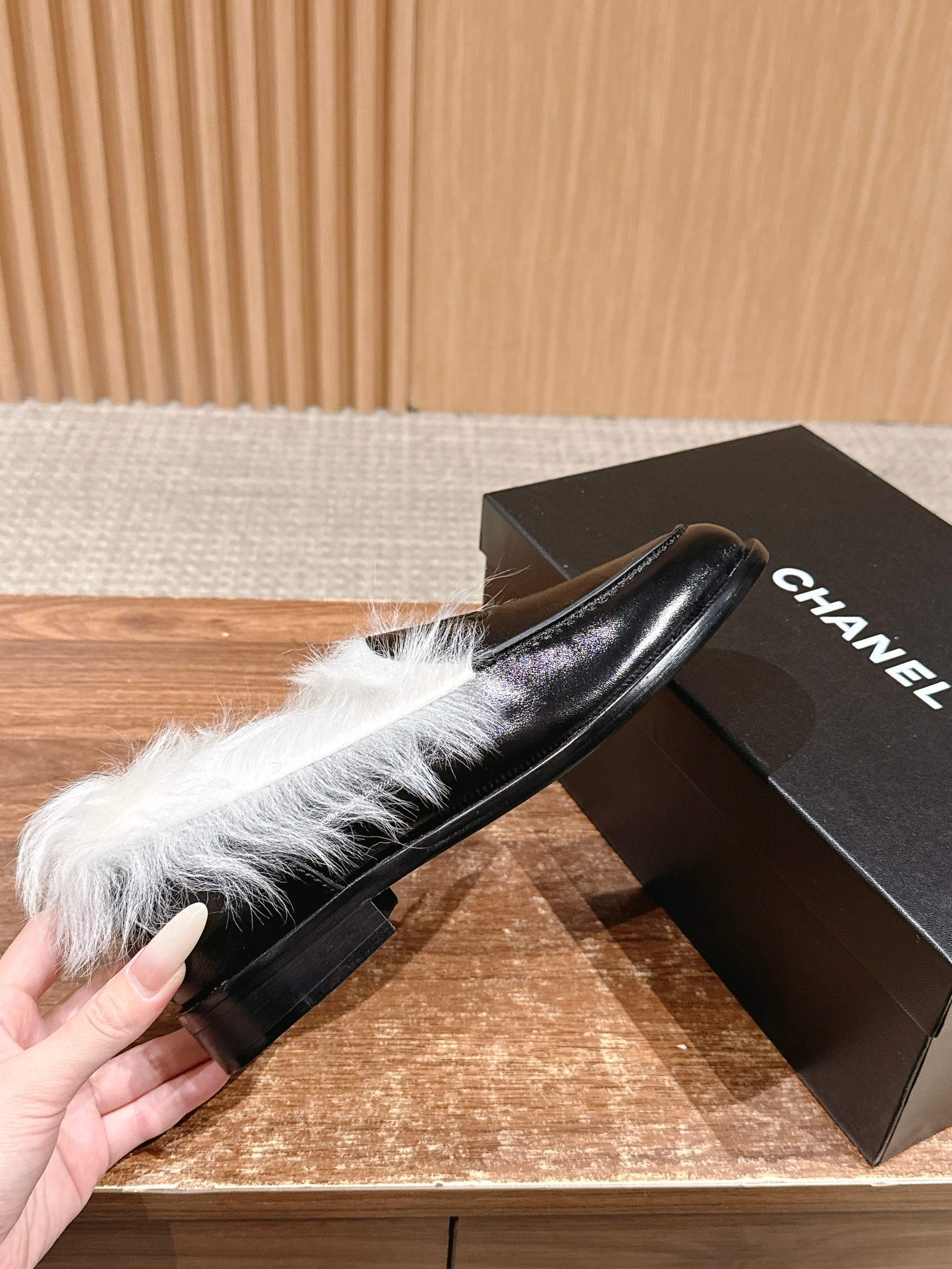 A pair of luxury black leather Chanel loafers with prominent white fur lining and CC logo on a wooden surface. - Hình 5