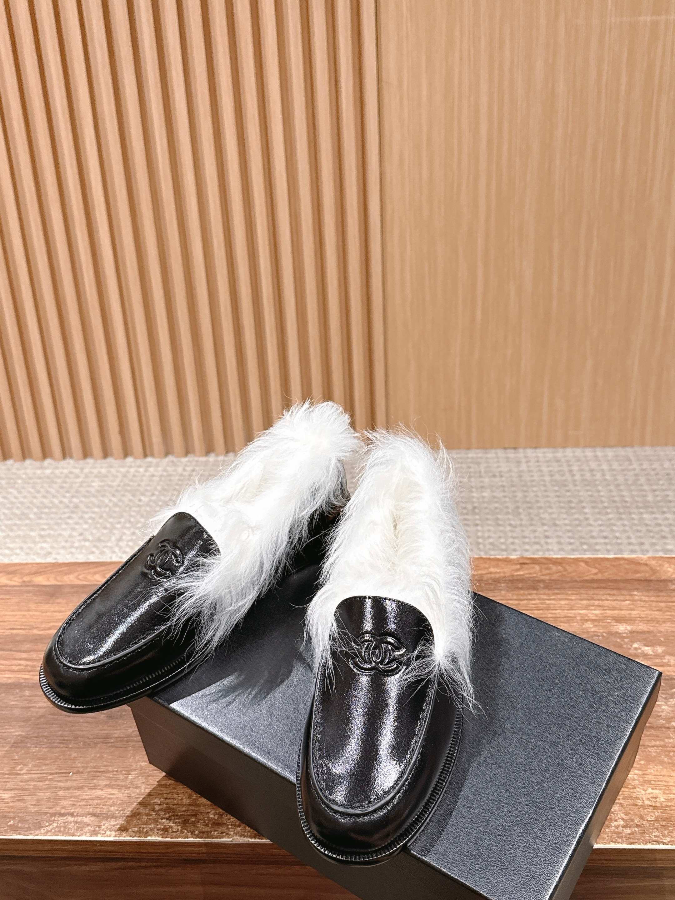 A pair of luxury black leather Chanel loafers with prominent white fur lining and CC logo on a wooden surface. - Hình 4