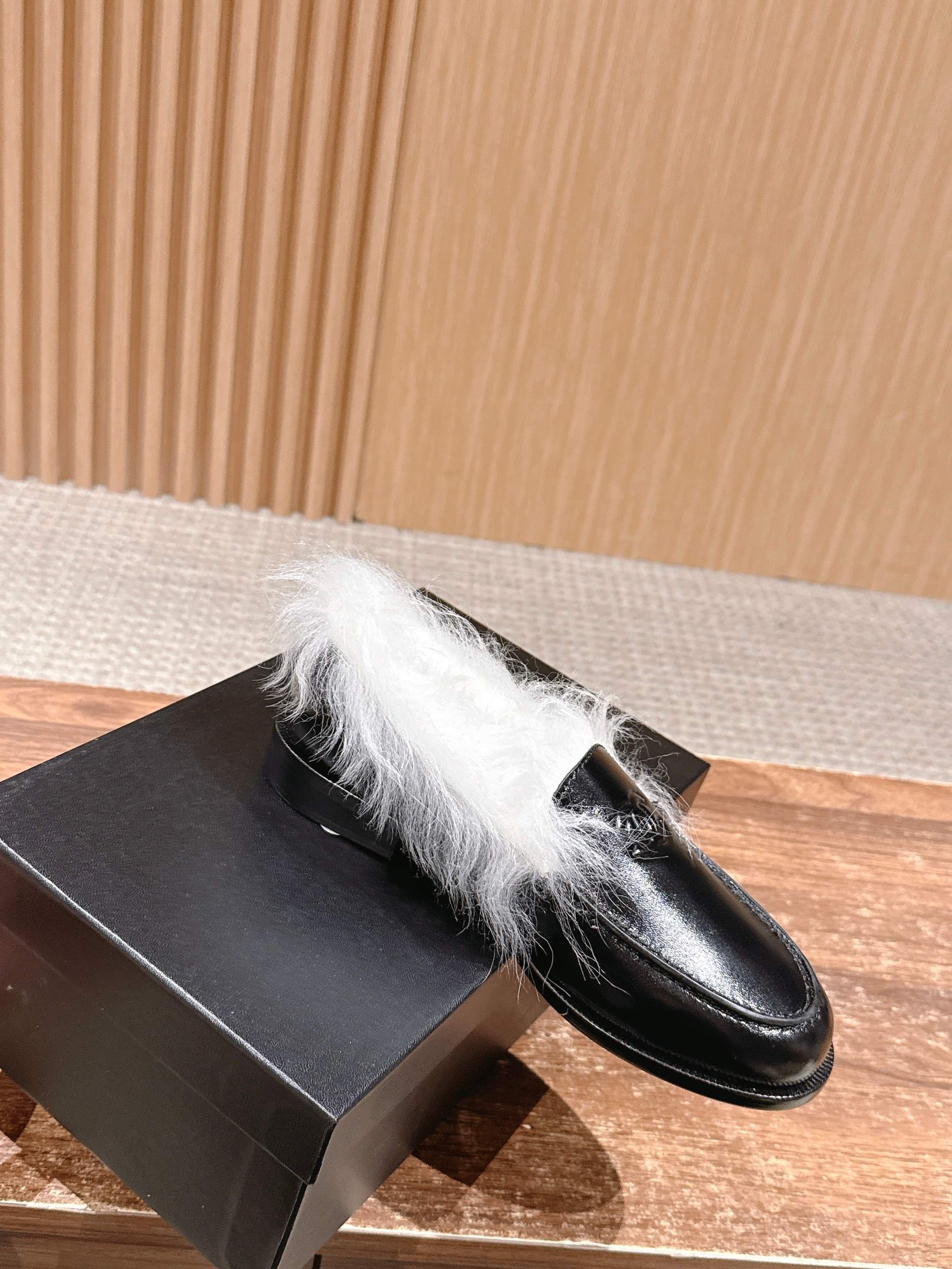 A pair of luxury black leather Chanel loafers with prominent white fur lining and CC logo on a wooden surface. - Hình 3