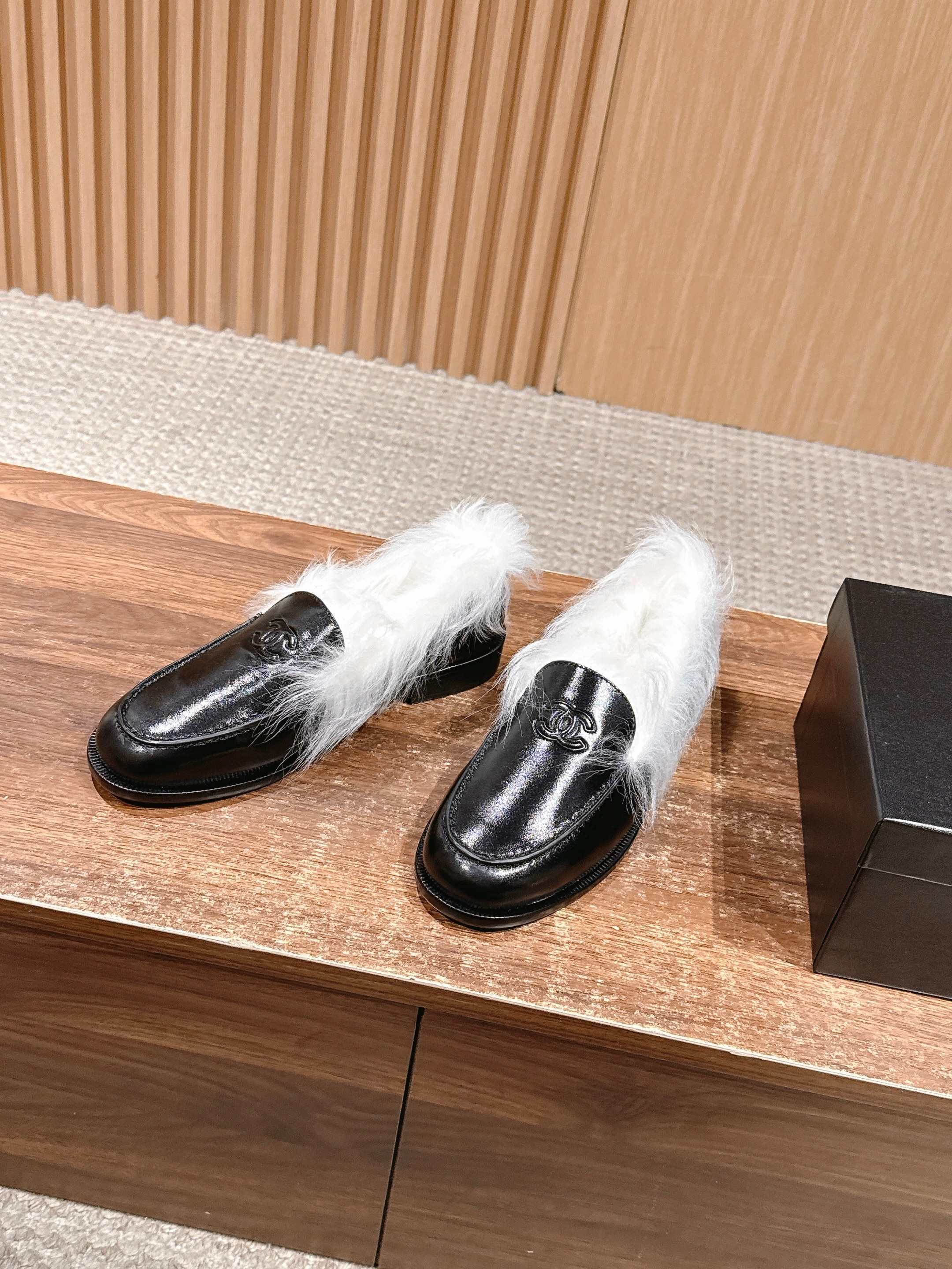 A pair of luxury black leather Chanel loafers with prominent white fur lining and CC logo on a wooden surface. - Hình 2