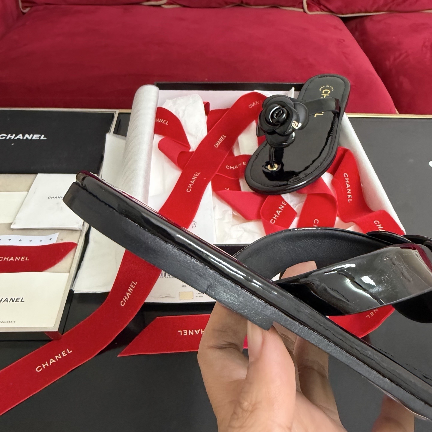 Authentic black Chanel thong sandals featuring a large 3D Camellia flower with a small gold CC logo, presented in a gift box with a red Chanel ribbon. - Hình 8