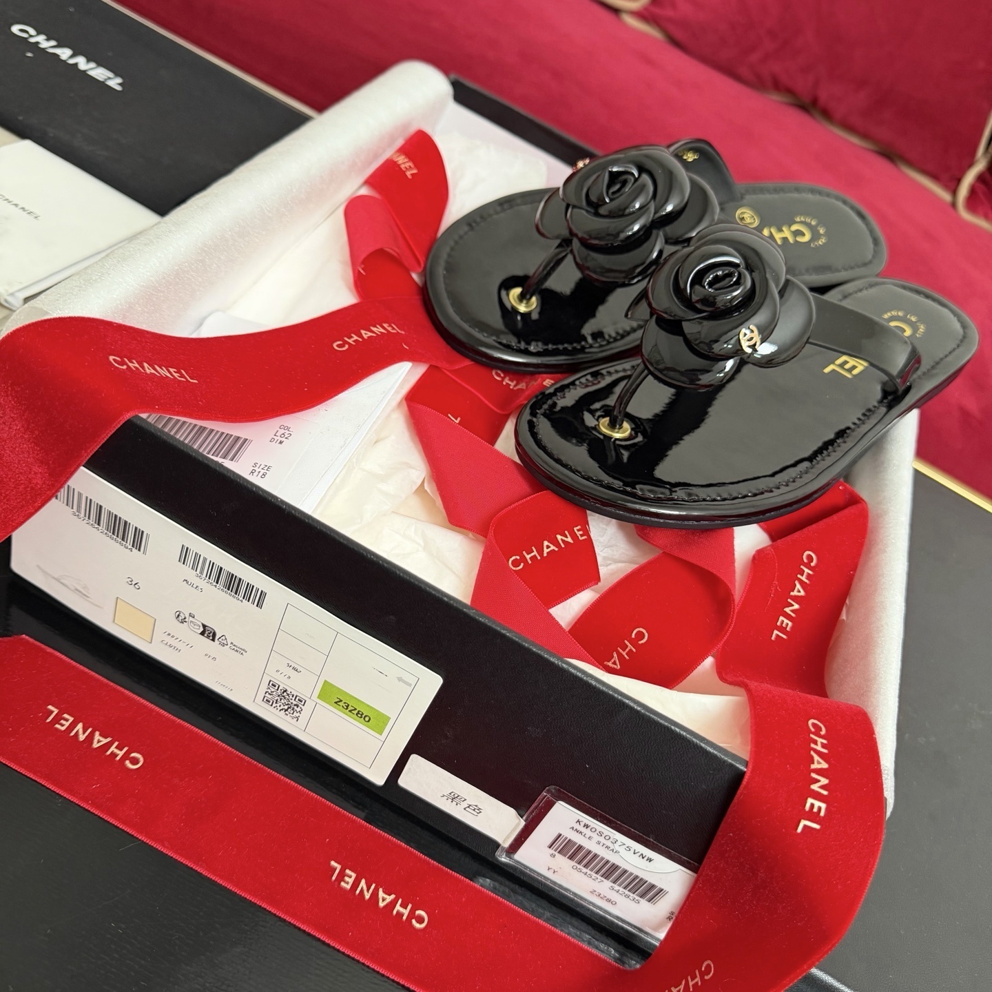 Authentic black Chanel thong sandals featuring a large 3D Camellia flower with a small gold CC logo, presented in a gift box with a red Chanel ribbon. - Hình 2