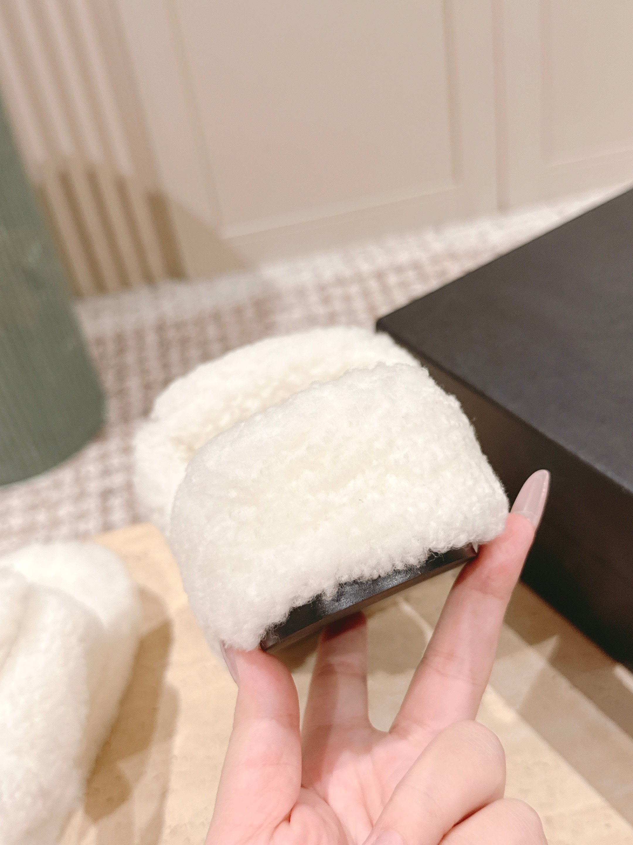 A pair of luxury Chanel white shearling slides featuring gold interlocking CC logos on the straps, displayed on a stone pedestal. - Hình 9