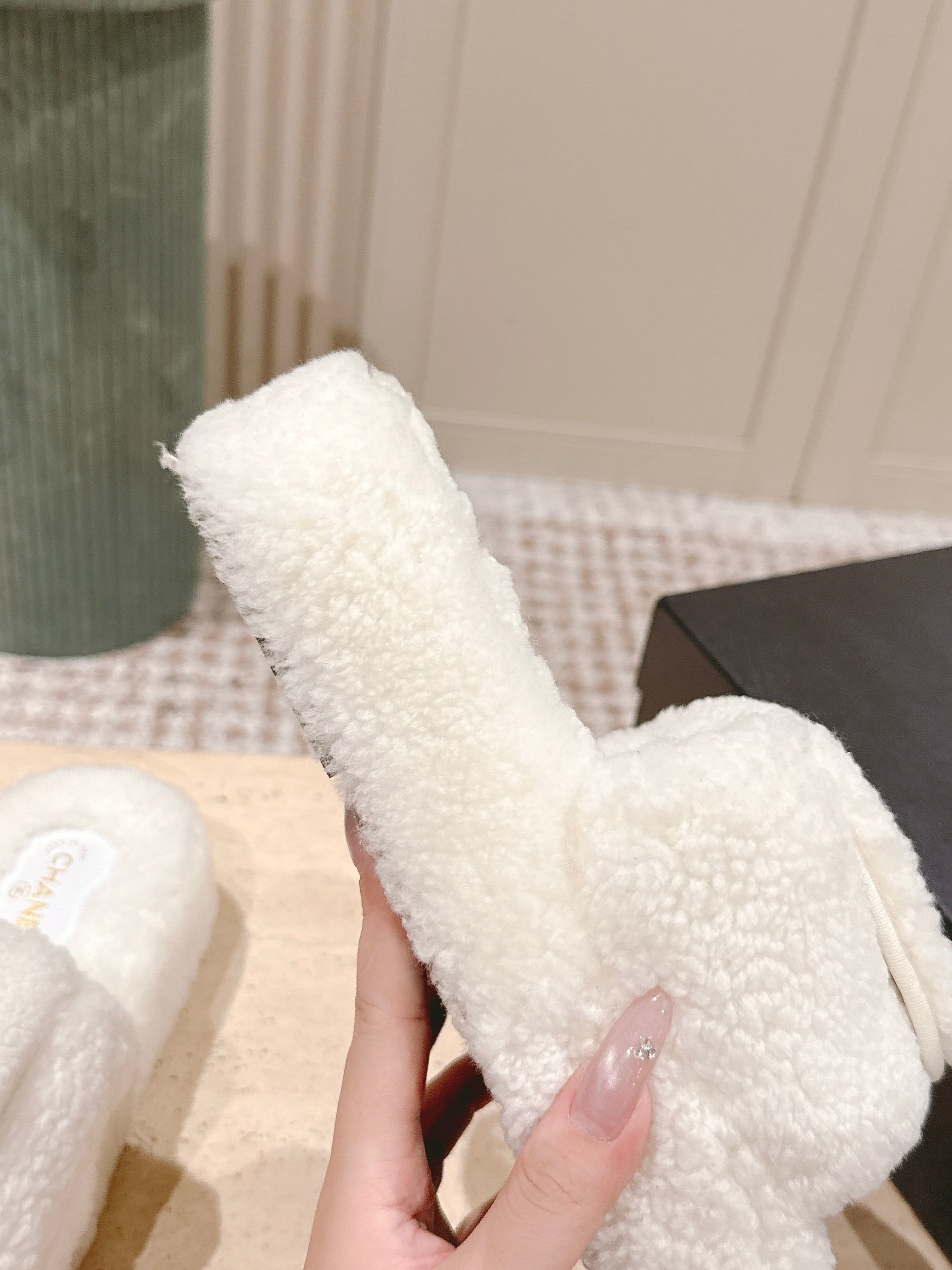 A pair of luxury Chanel white shearling slides featuring gold interlocking CC logos on the straps, displayed on a stone pedestal. - Hình 8