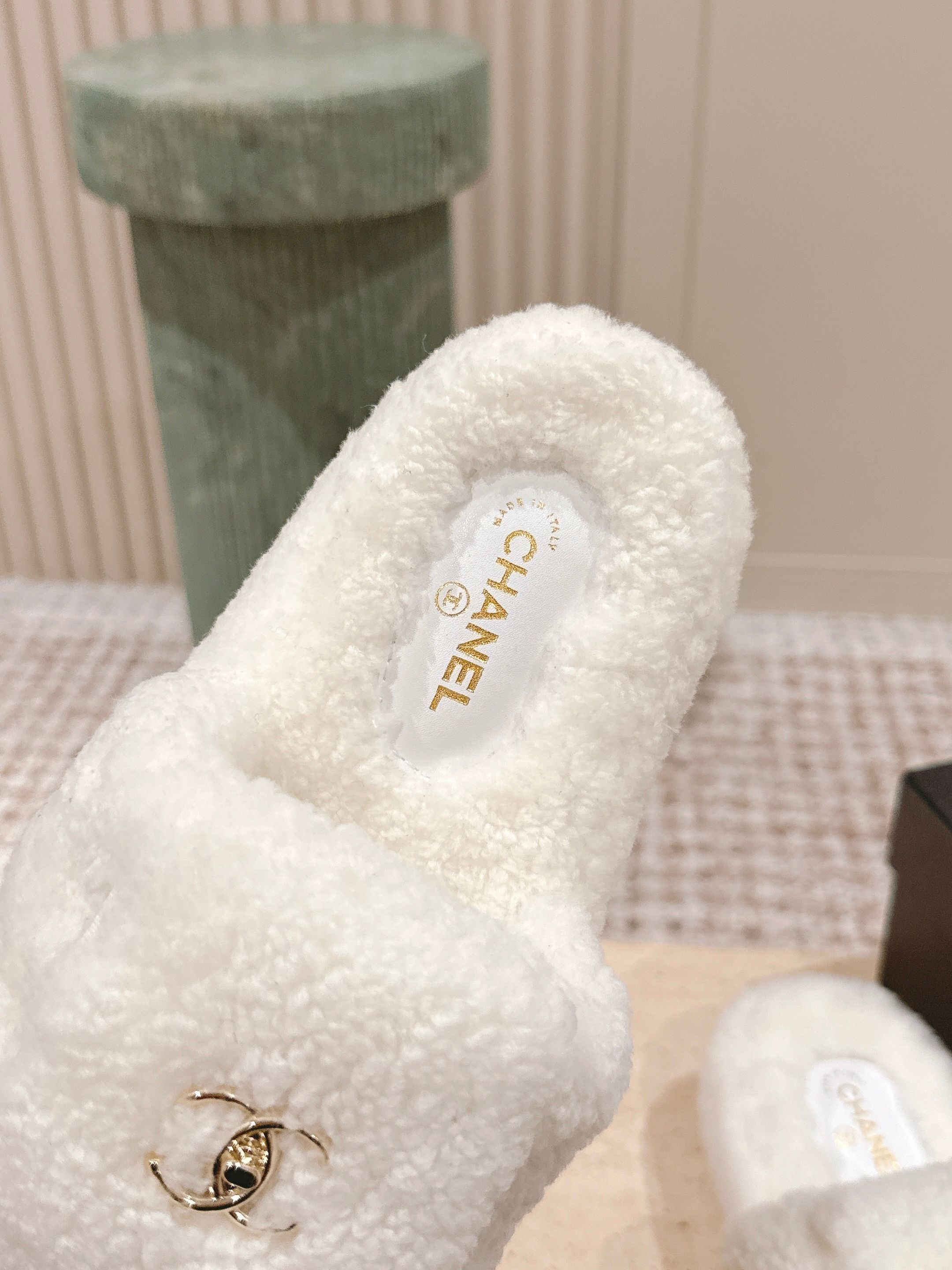 A pair of luxury Chanel white shearling slides featuring gold interlocking CC logos on the straps, displayed on a stone pedestal. - Hình 7