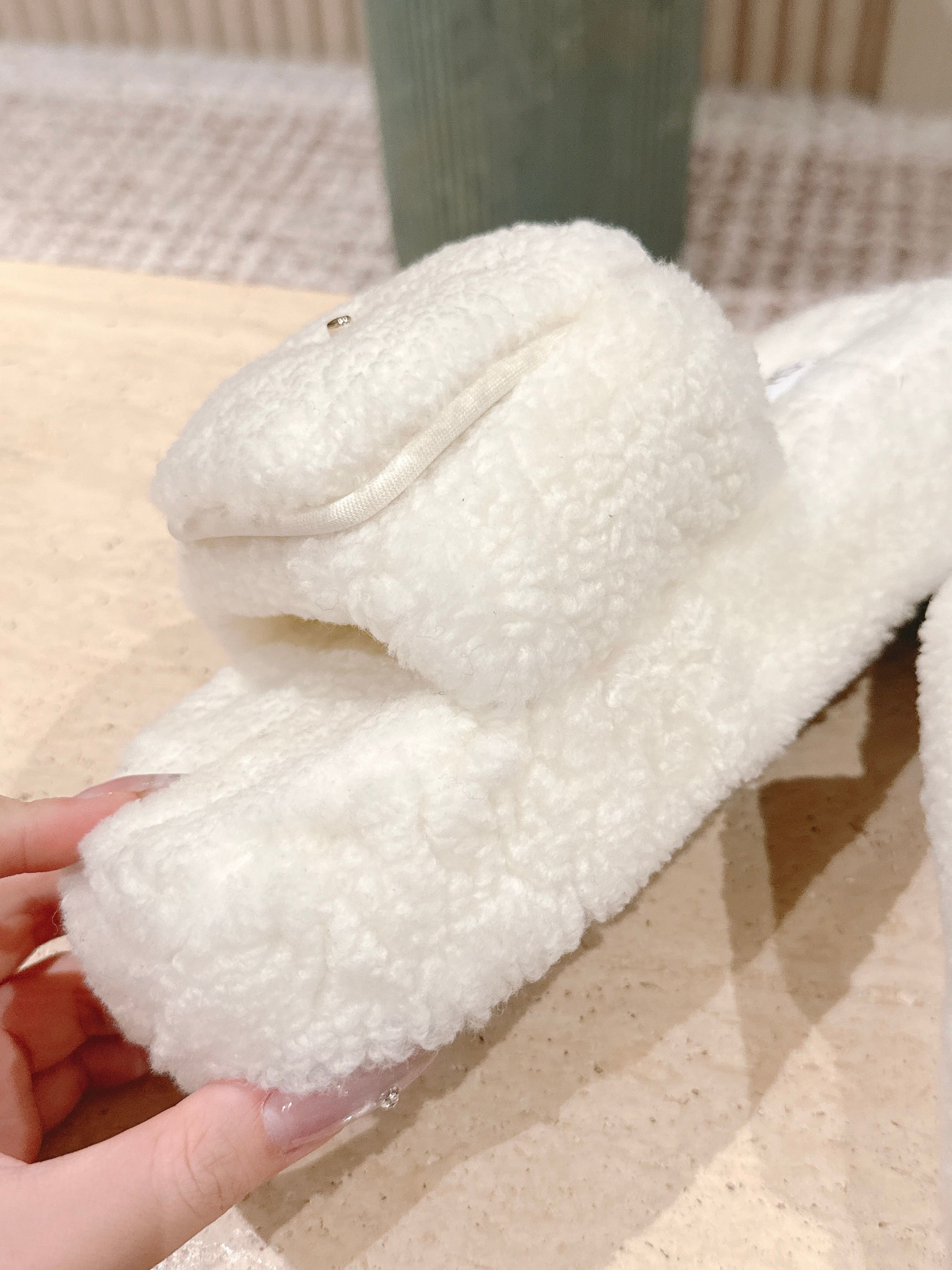 A pair of luxury Chanel white shearling slides featuring gold interlocking CC logos on the straps, displayed on a stone pedestal. - Hình 6