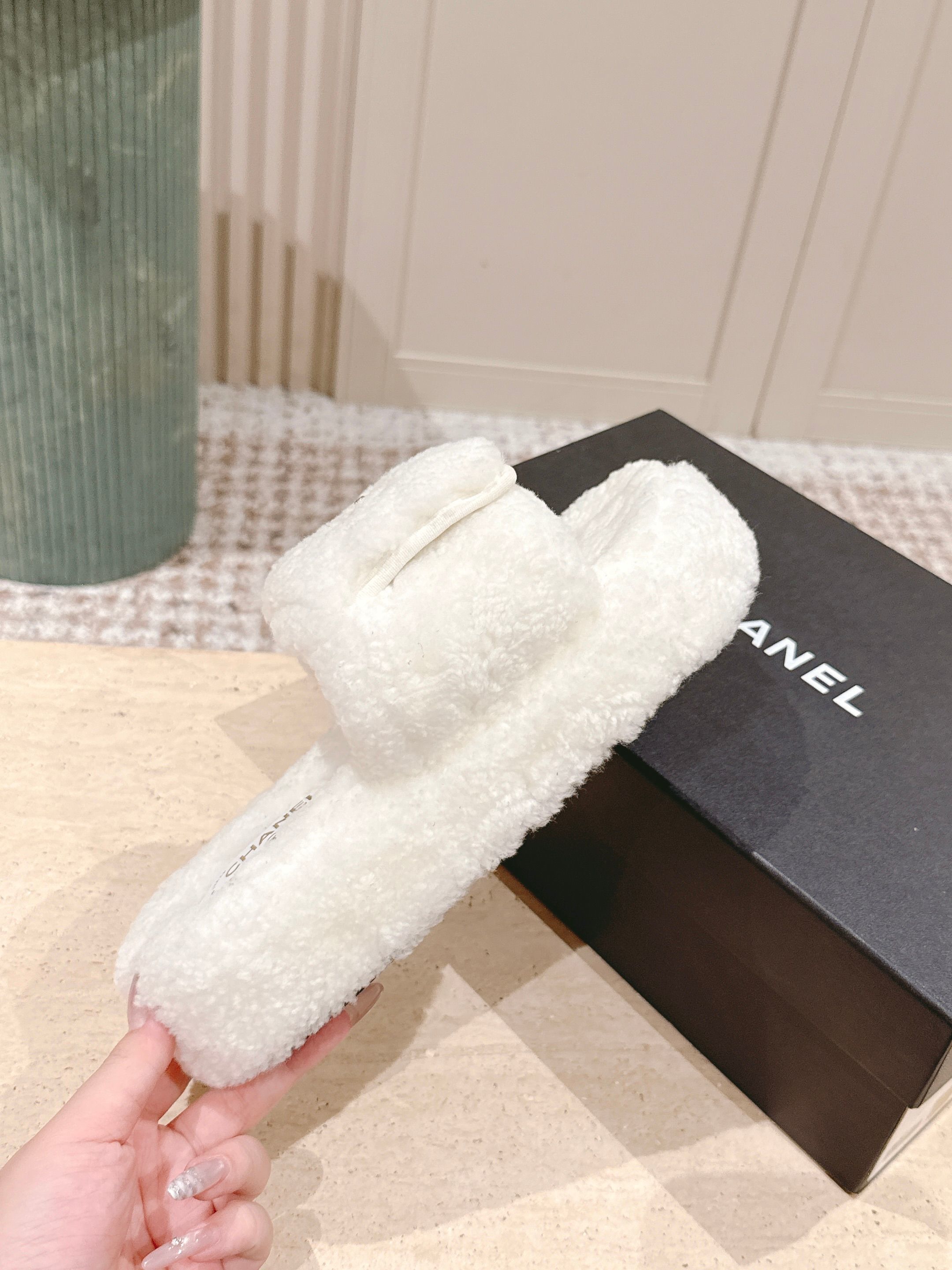 A pair of luxury Chanel white shearling slides featuring gold interlocking CC logos on the straps, displayed on a stone pedestal. - Hình 5