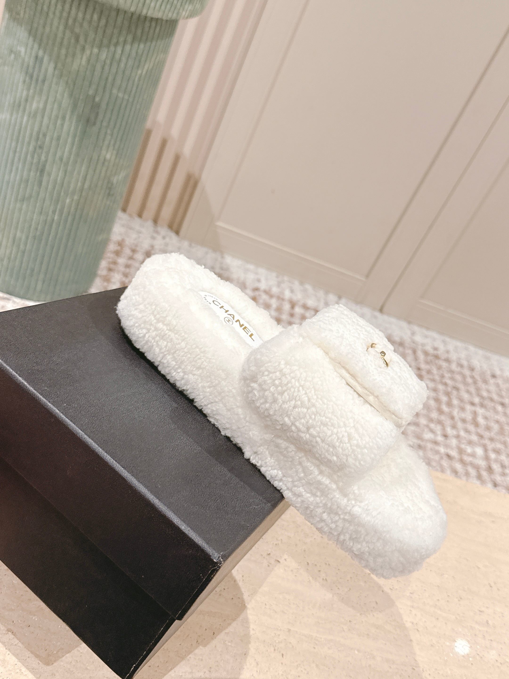A pair of luxury Chanel white shearling slides featuring gold interlocking CC logos on the straps, displayed on a stone pedestal. - Hình 4