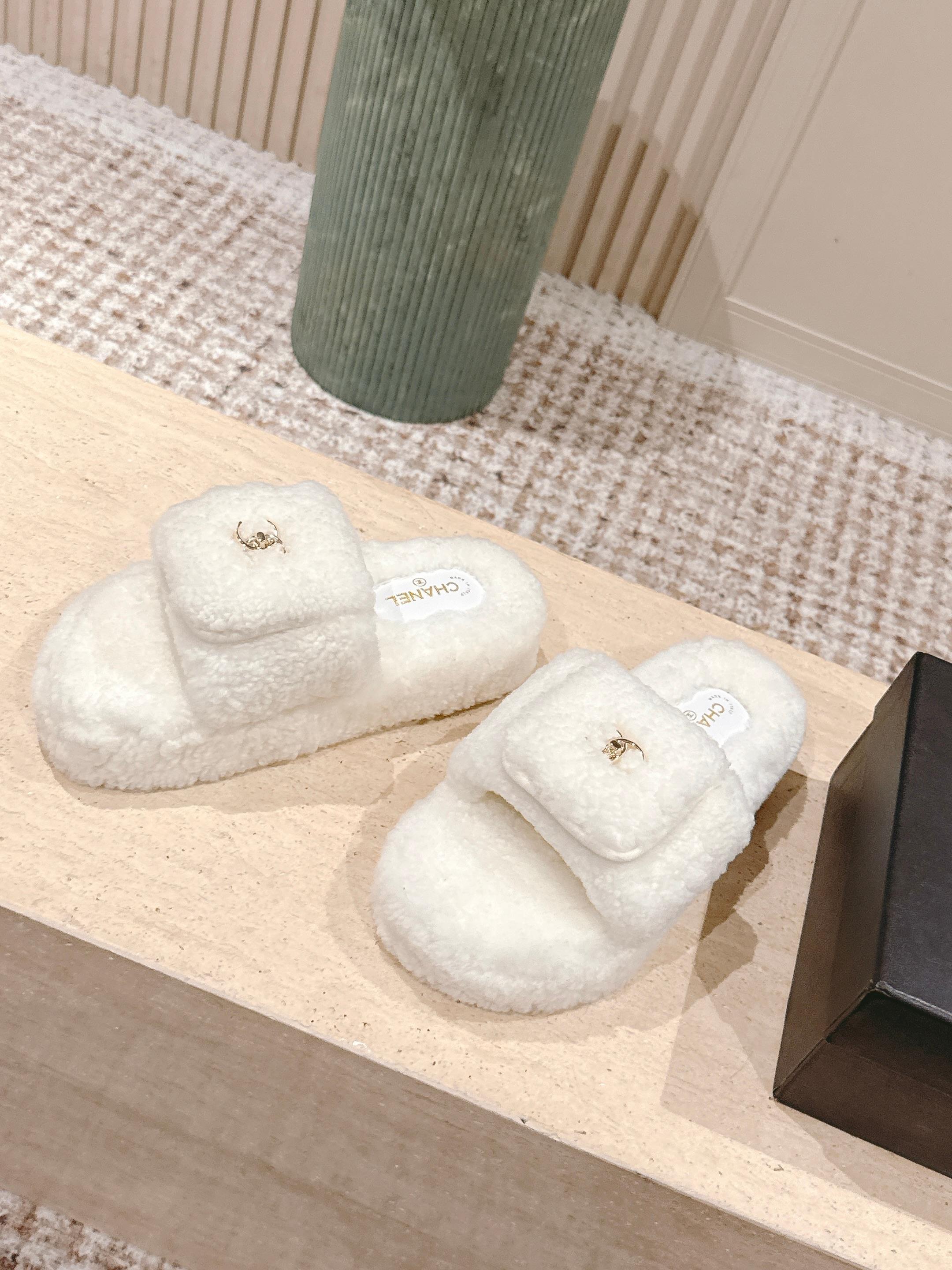 A pair of luxury Chanel white shearling slides featuring gold interlocking CC logos on the straps, displayed on a stone pedestal. - Hình 3