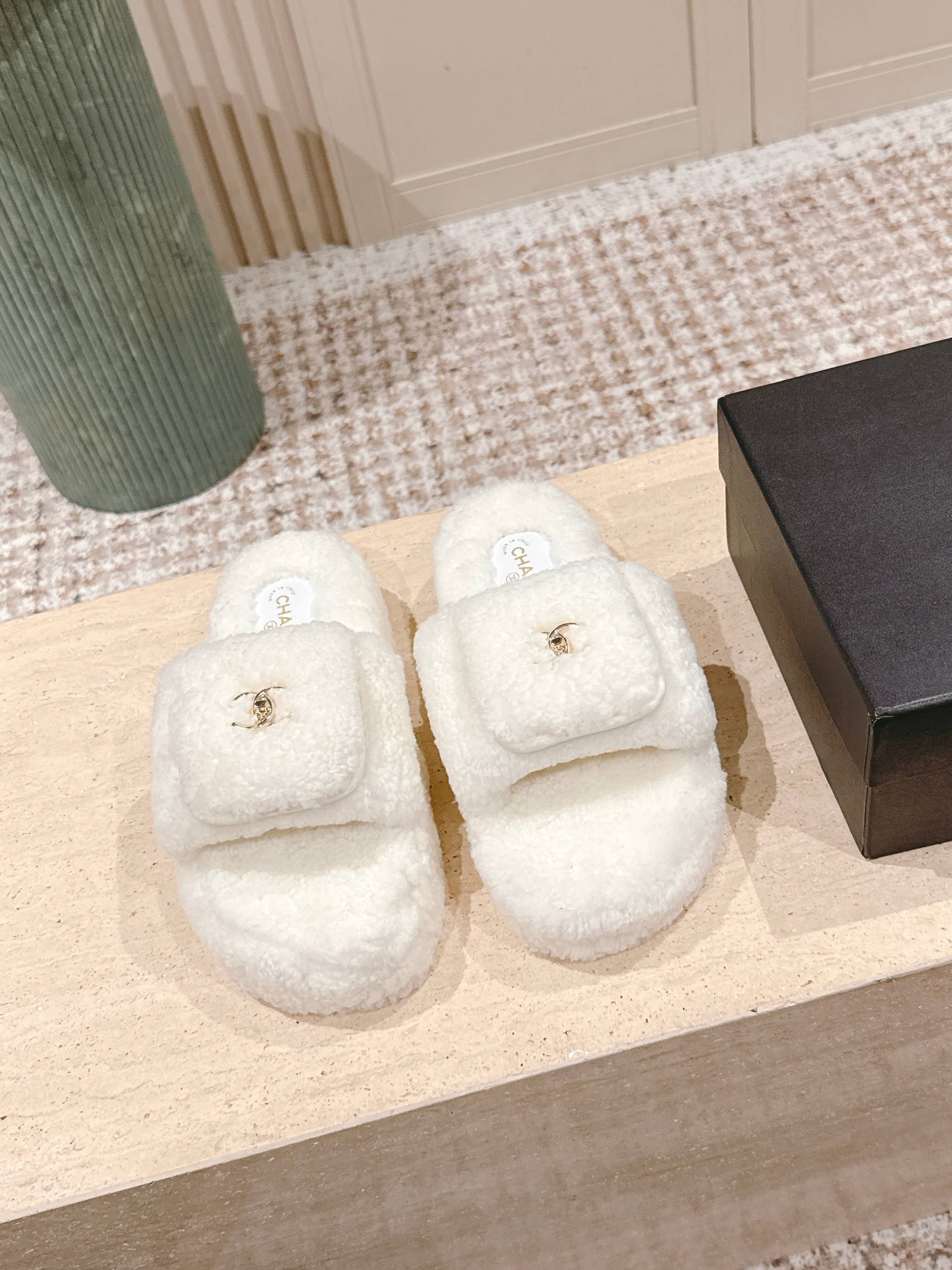 A pair of luxury Chanel white shearling slides featuring gold interlocking CC logos on the straps, displayed on a stone pedestal. - Hình 2
