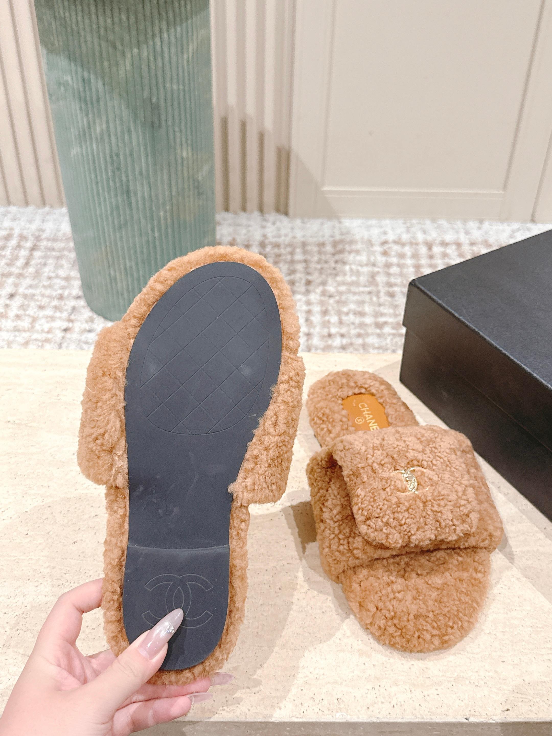 A pair of Chanel camel brown shearling slides featuring the gold CC logo on the strap, displayed on a marble countertop next to a black shoe box. - Hình 9