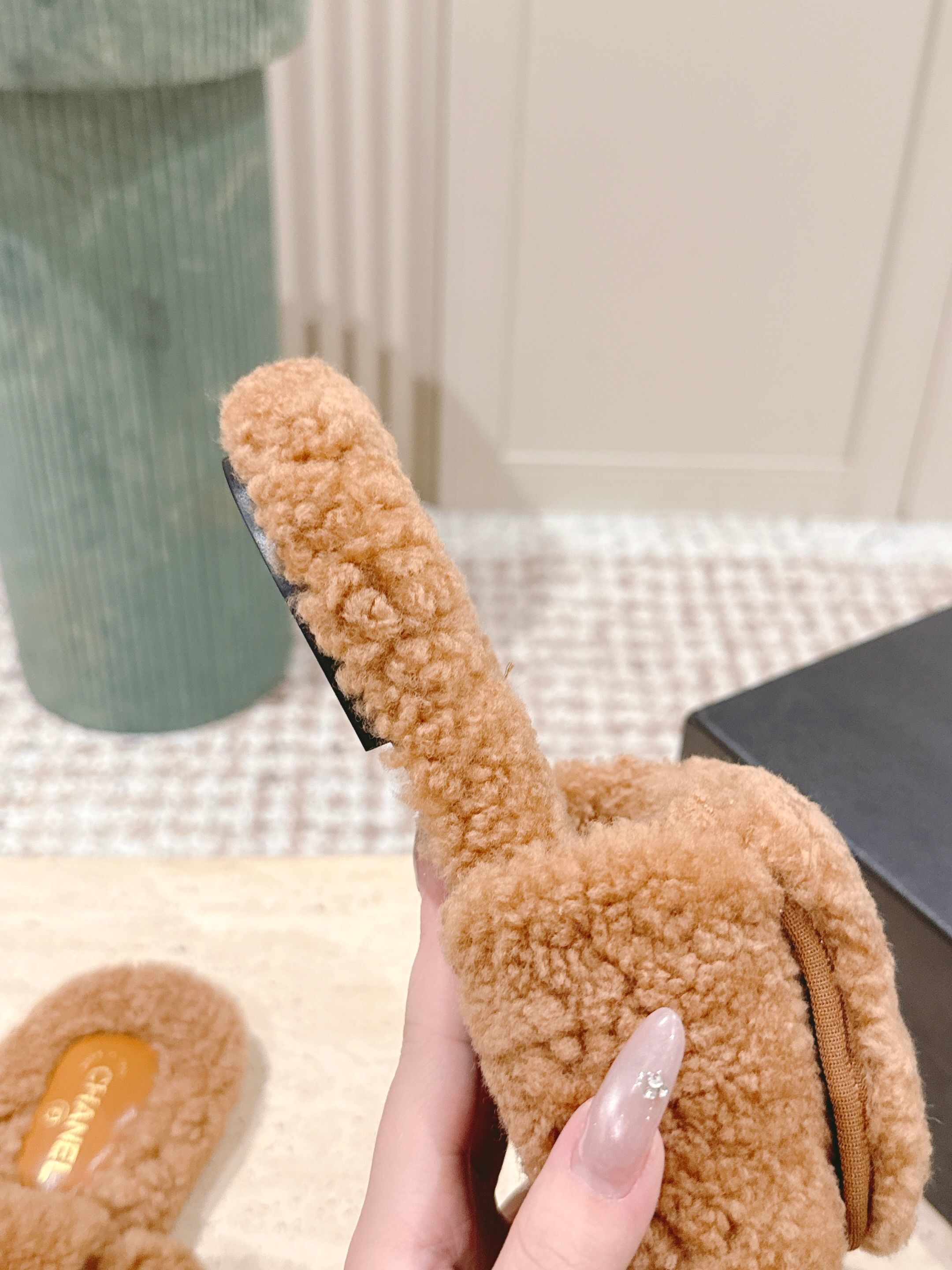 A pair of Chanel camel brown shearling slides featuring the gold CC logo on the strap, displayed on a marble countertop next to a black shoe box. - Hình 8