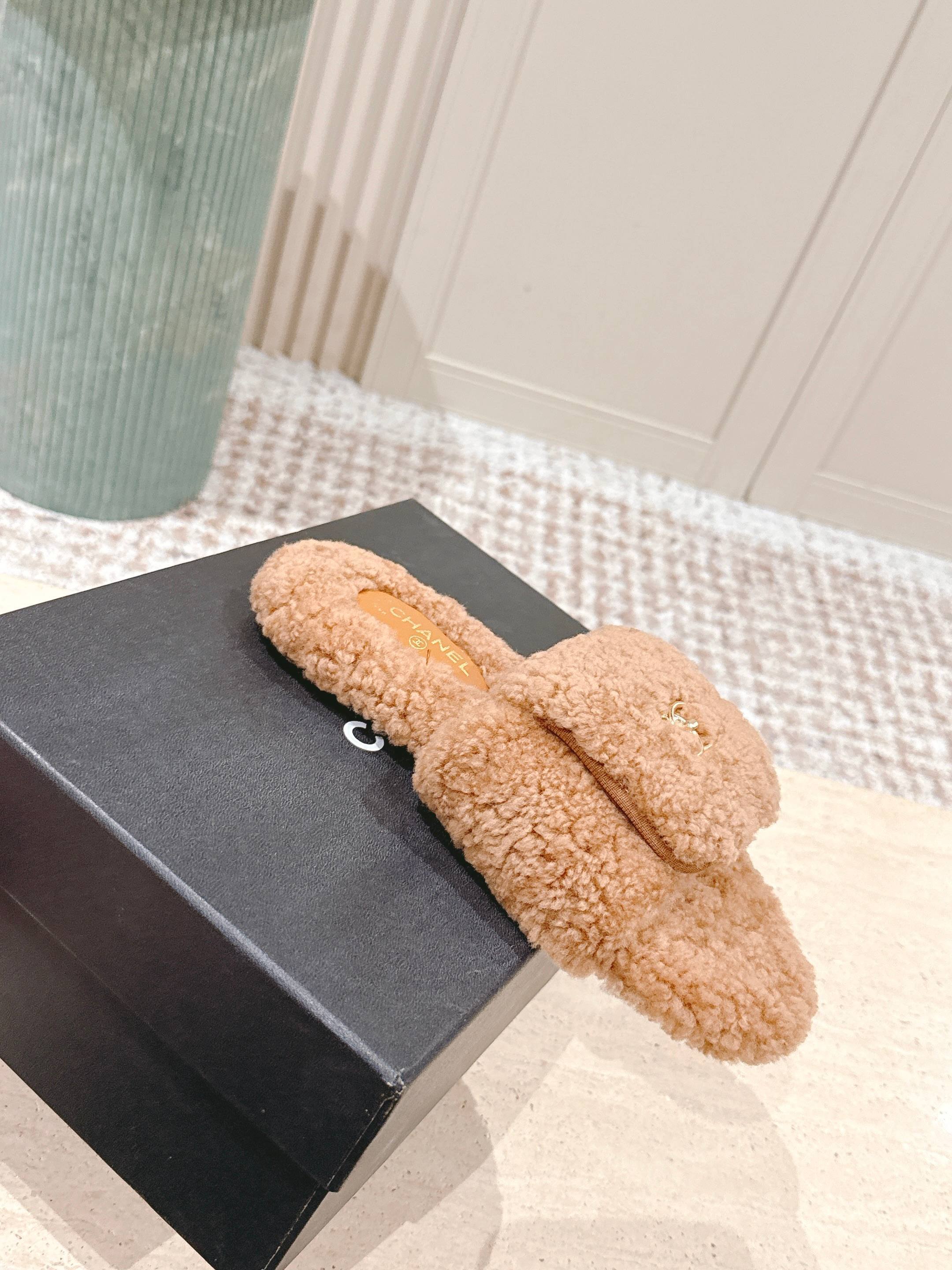 A pair of Chanel camel brown shearling slides featuring the gold CC logo on the strap, displayed on a marble countertop next to a black shoe box. - Hình 4