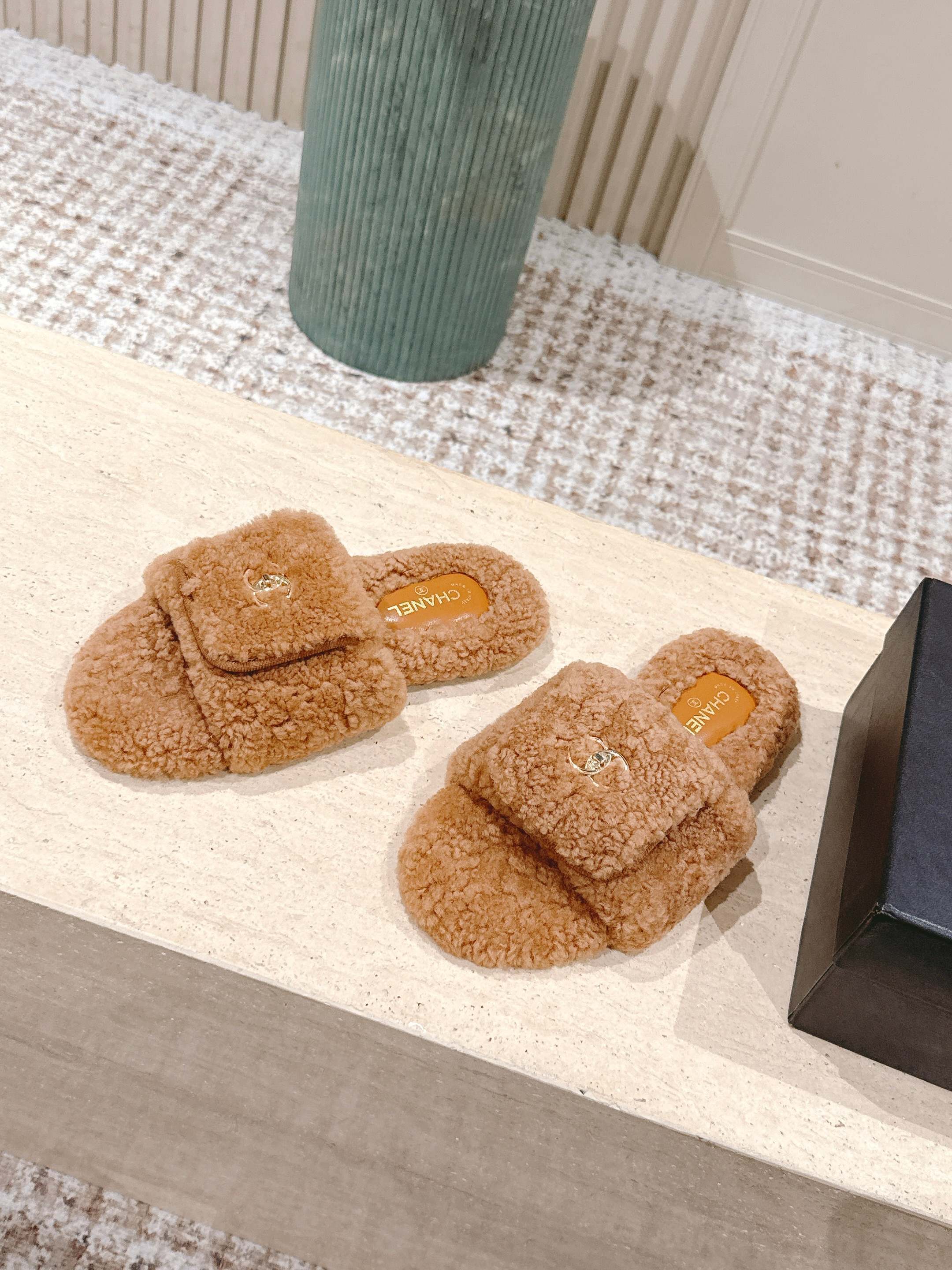A pair of Chanel camel brown shearling slides featuring the gold CC logo on the strap, displayed on a marble countertop next to a black shoe box. - Hình 3