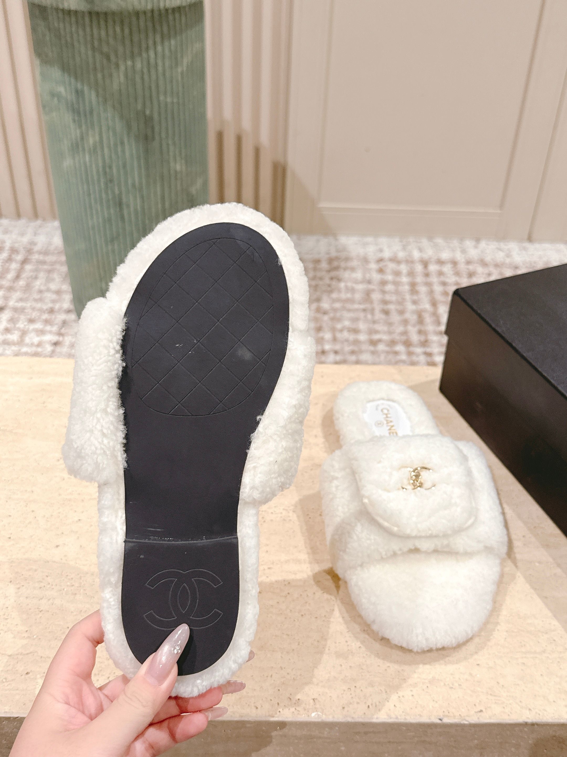 A pair of white Chanel shearling slides with gold CC logo displayed on a stone pedestal - Hình 9