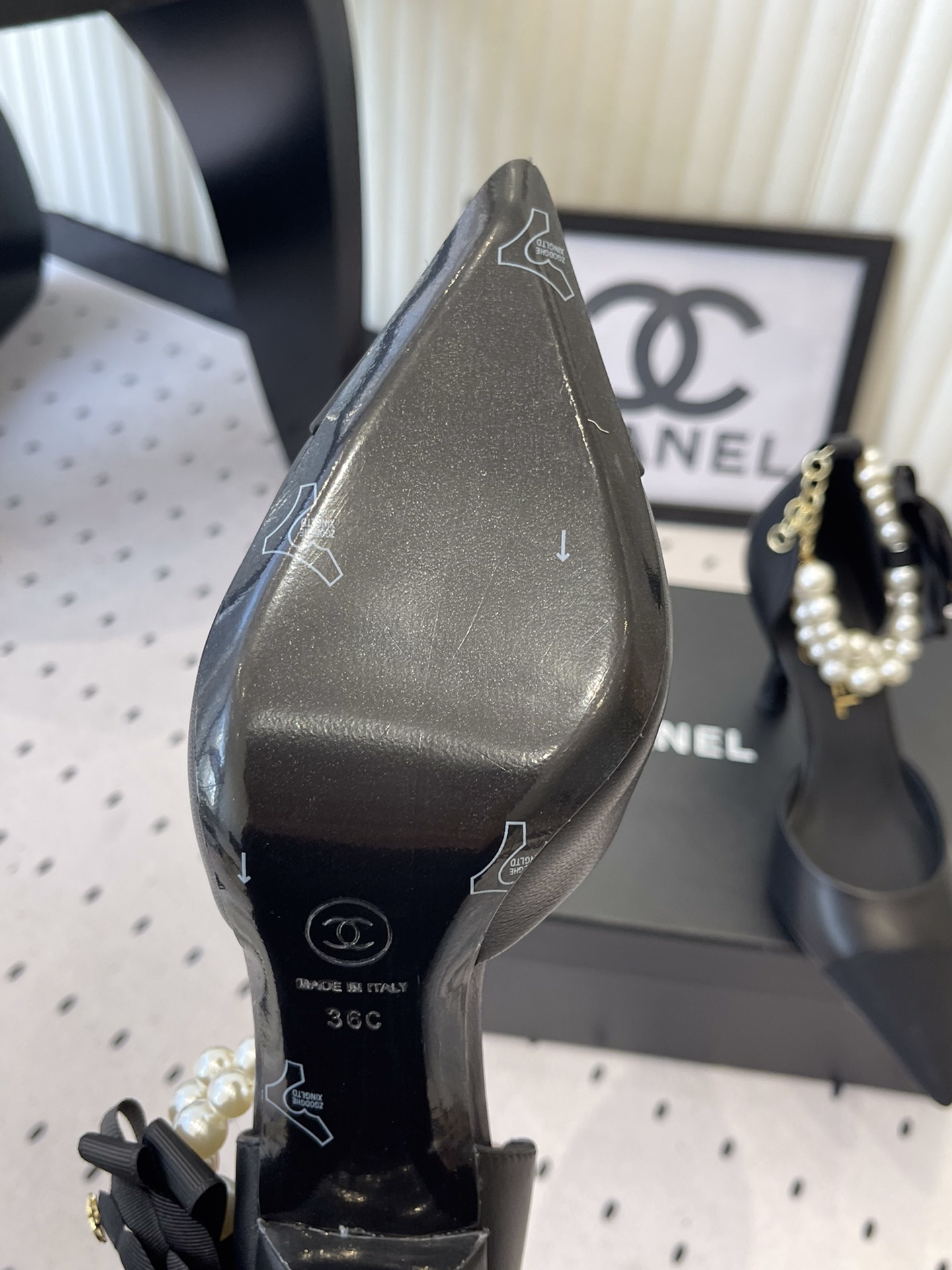 Pair of Chanel black leather pointed-toe pumps featuring black camellia flowers and pearl ankle straps displayed on a black Chanel box. - Hình 9