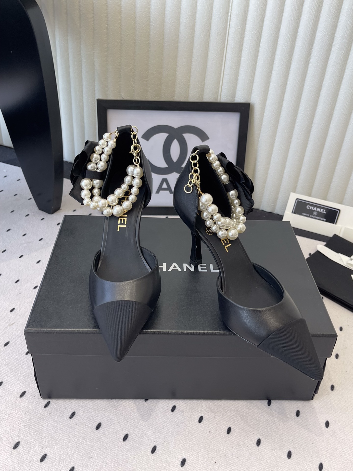 Pair of Chanel black leather pointed-toe pumps featuring black camellia flowers and pearl ankle straps displayed on a black Chanel box. - Hình 8