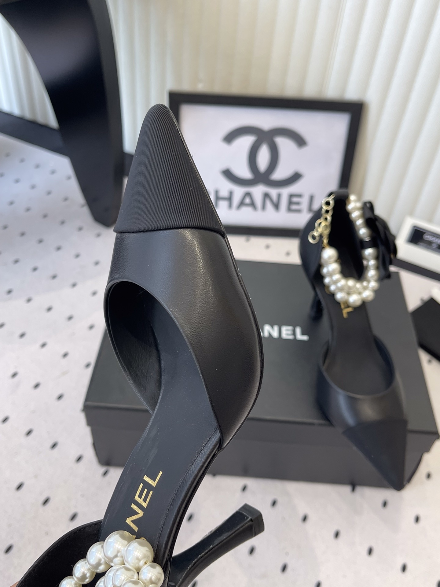 Pair of Chanel black leather pointed-toe pumps featuring black camellia flowers and pearl ankle straps displayed on a black Chanel box. - Hình 7