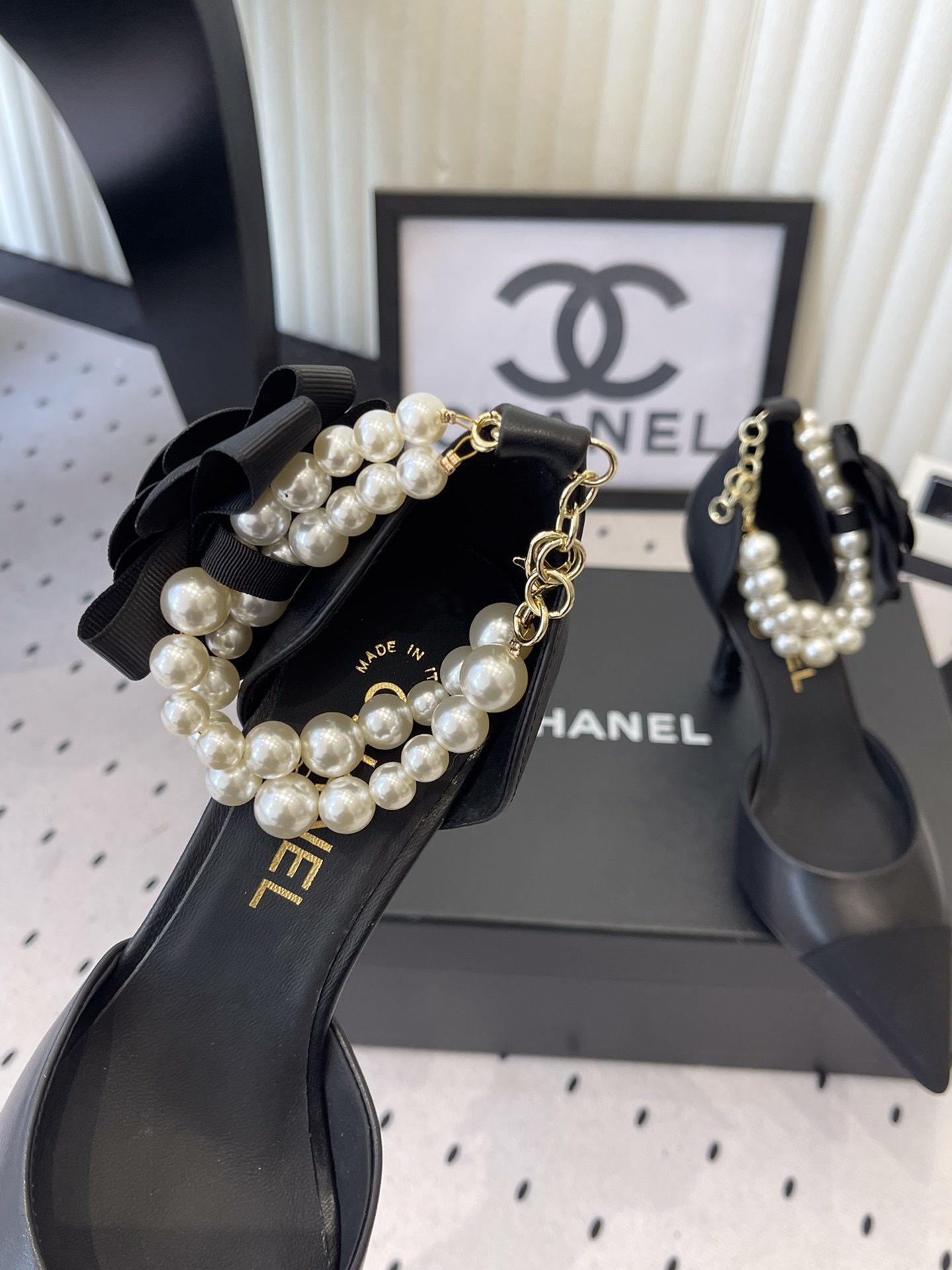 Pair of Chanel black leather pointed-toe pumps featuring black camellia flowers and pearl ankle straps displayed on a black Chanel box. - Hình 6