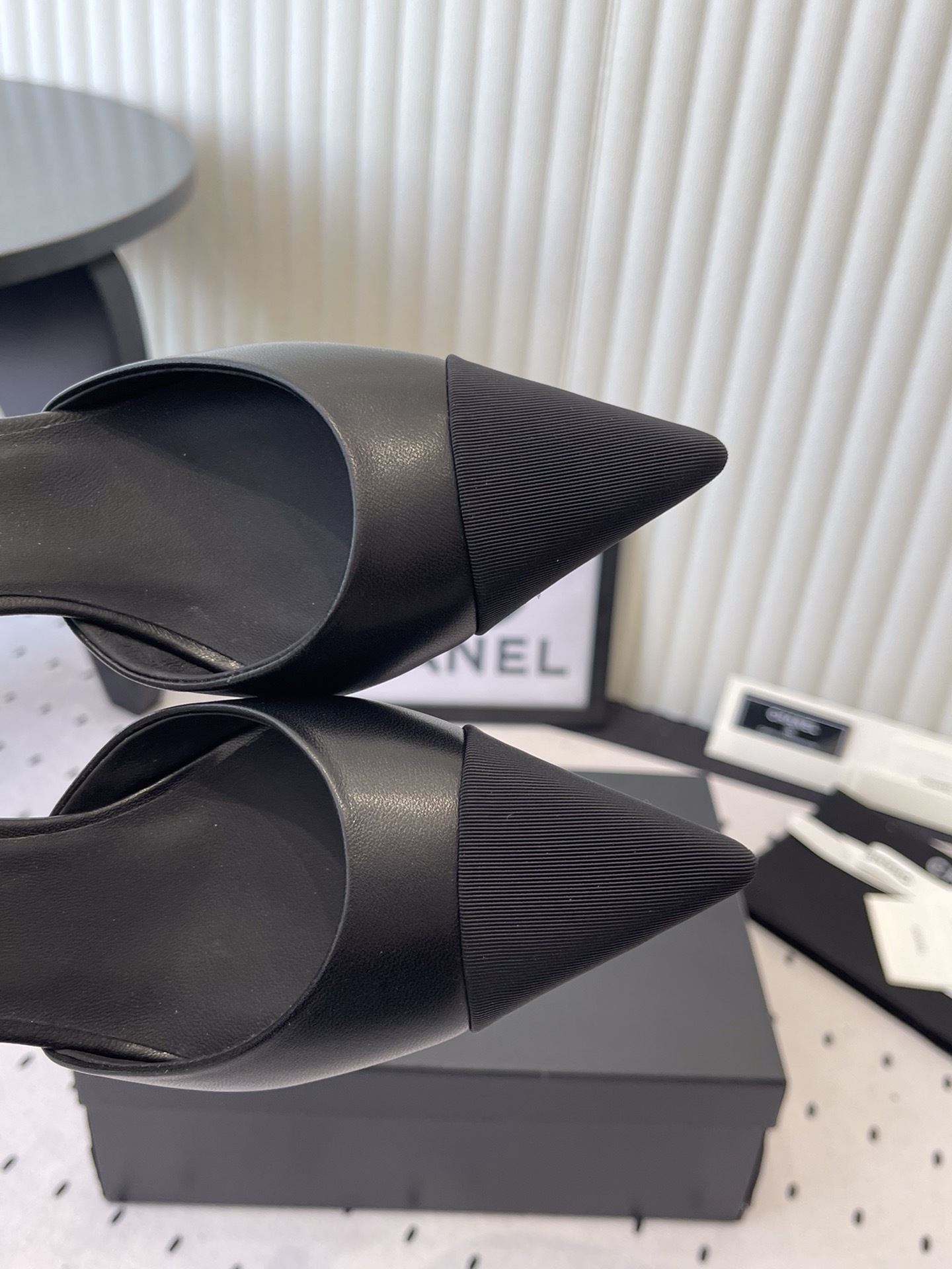 Pair of Chanel black leather pointed-toe pumps featuring black camellia flowers and pearl ankle straps displayed on a black Chanel box. - Hình 4