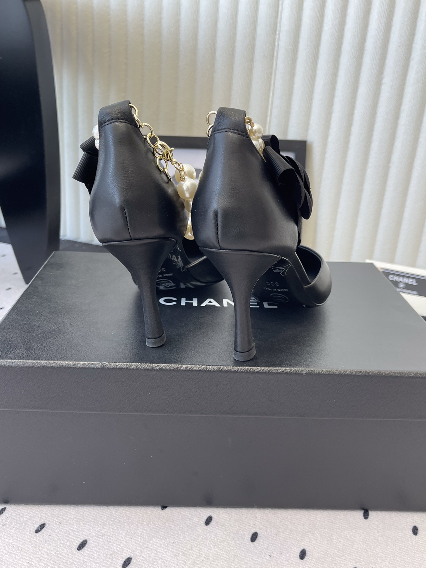 Pair of Chanel black leather pointed-toe pumps featuring black camellia flowers and pearl ankle straps displayed on a black Chanel box. - Hình 3