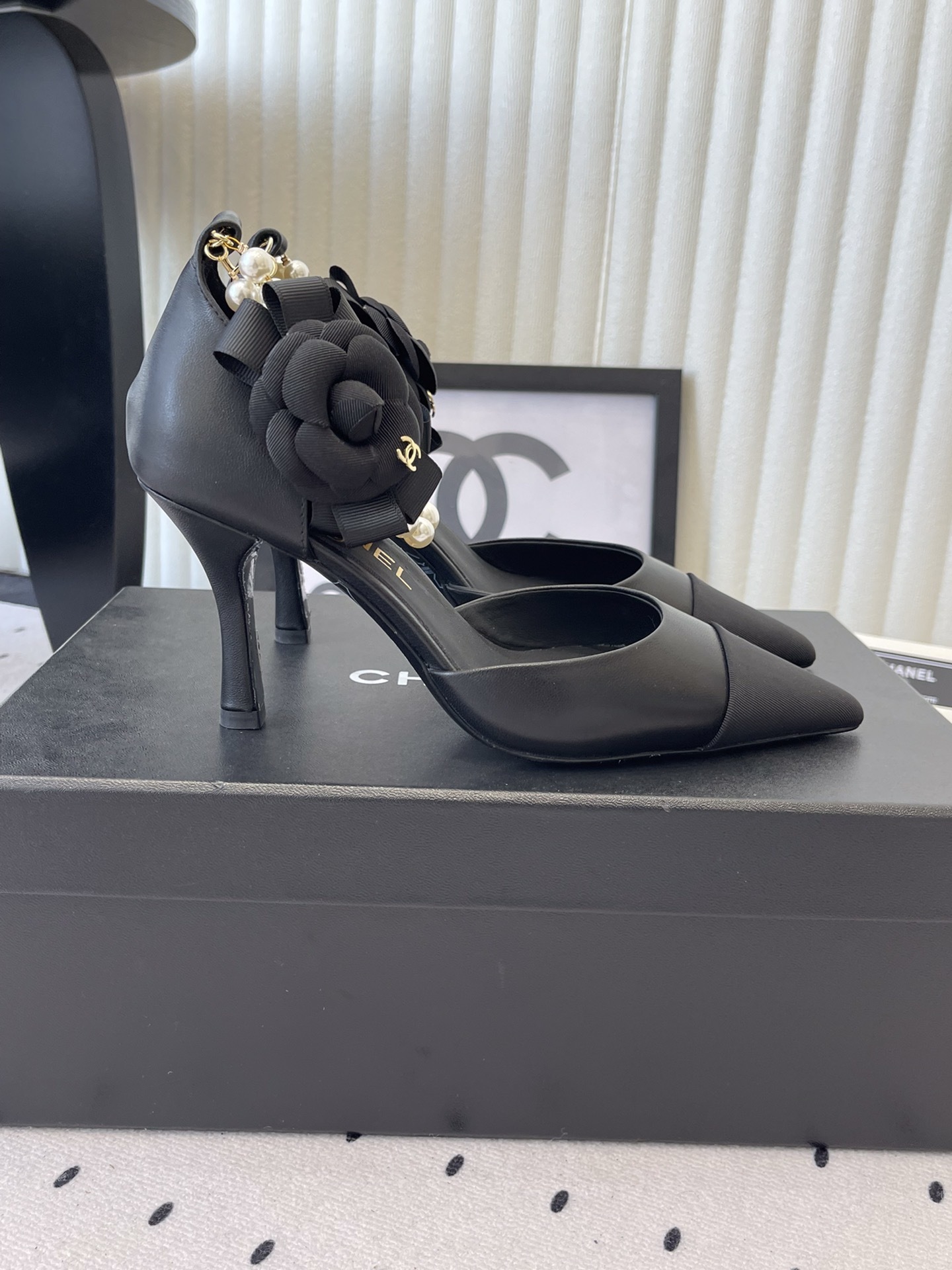 Pair of Chanel black leather pointed-toe pumps featuring black camellia flowers and pearl ankle straps displayed on a black Chanel box. - Hình 2