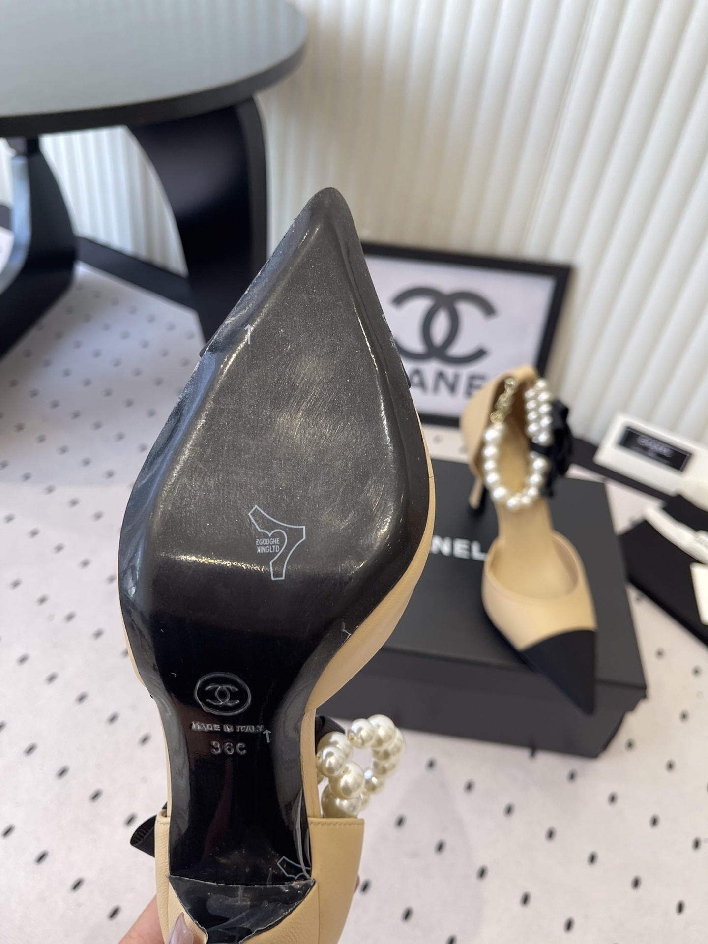 Luxury beige and black two-tone pointed-toe pumps with pearl ankle strap and black camellia flower detail on a designer box. - Hình 9