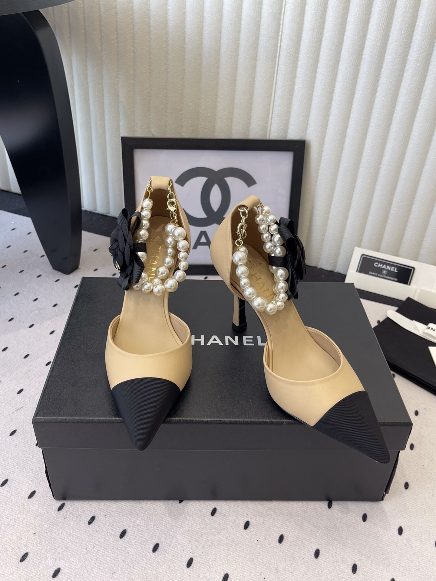 Luxury beige and black two-tone pointed-toe pumps with pearl ankle strap and black camellia flower detail on a designer box. - Hình 8