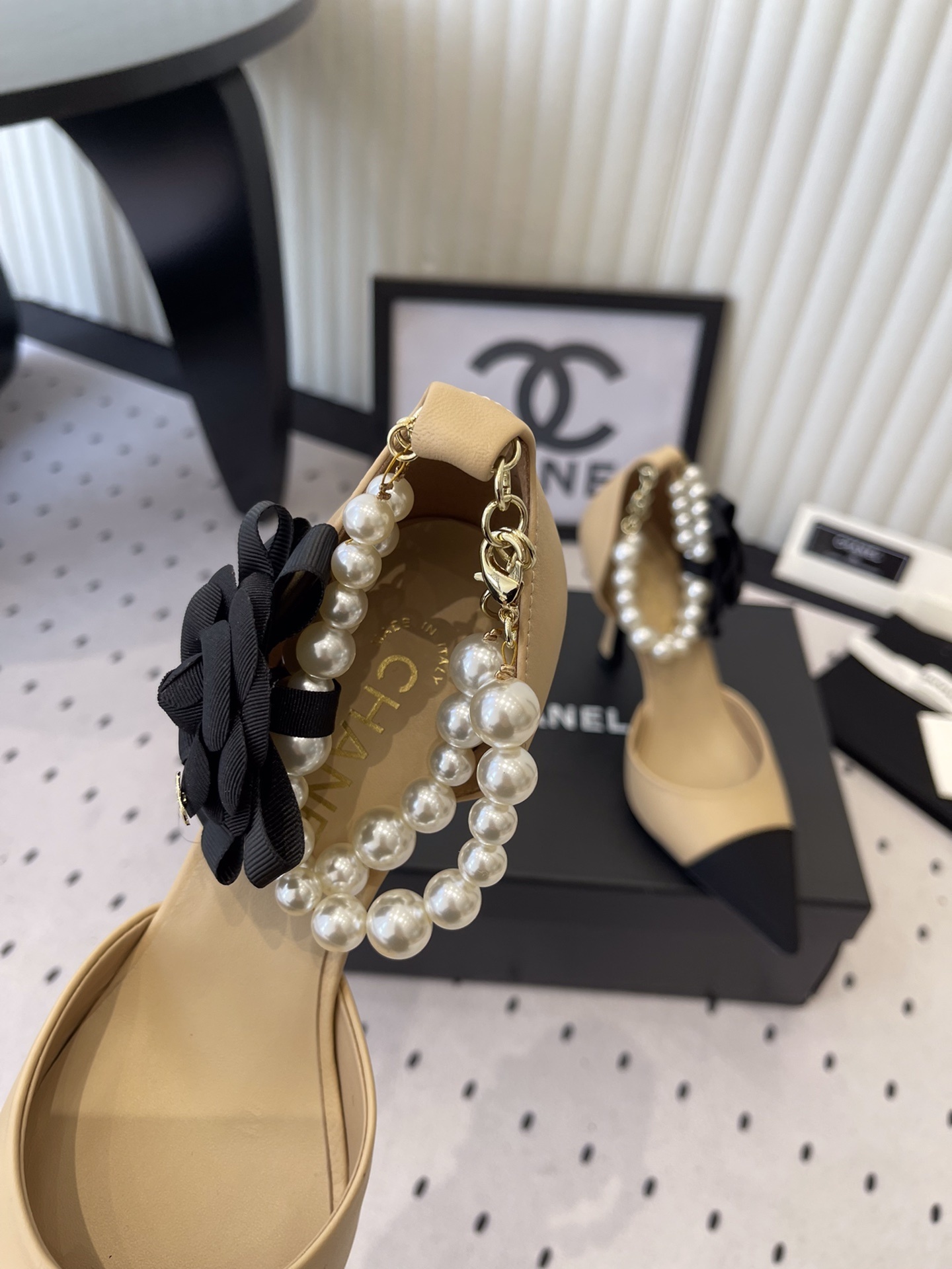 Luxury beige and black two-tone pointed-toe pumps with pearl ankle strap and black camellia flower detail on a designer box. - Hình 6