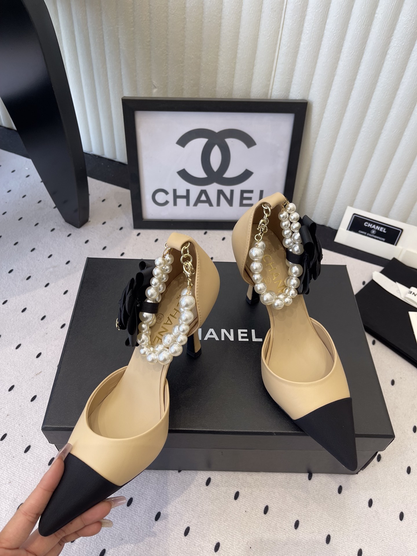 Luxury beige and black two-tone pointed-toe pumps with pearl ankle strap and black camellia flower detail on a designer box. - Hình 5