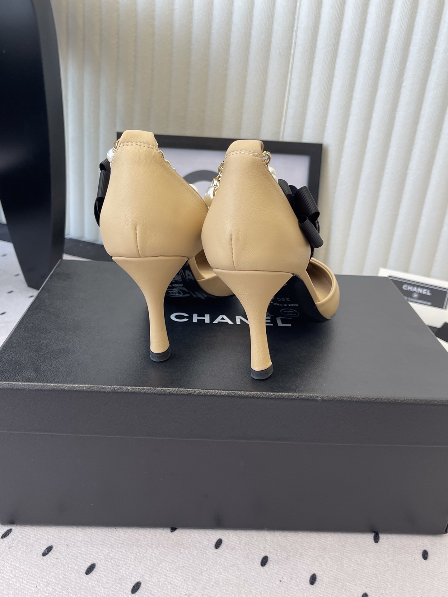 Luxury beige and black two-tone pointed-toe pumps with pearl ankle strap and black camellia flower detail on a designer box. - Hình 3