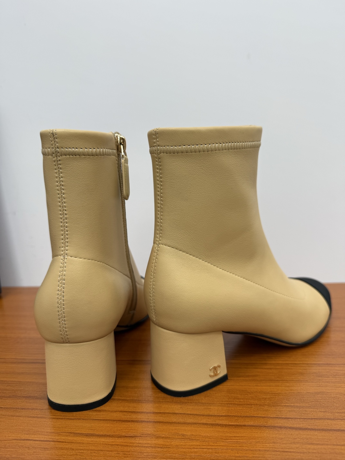 A pair of elegant beige leather-look ankle boots with a black cap toe and a sturdy block heel on a wooden surface. - Hình 8