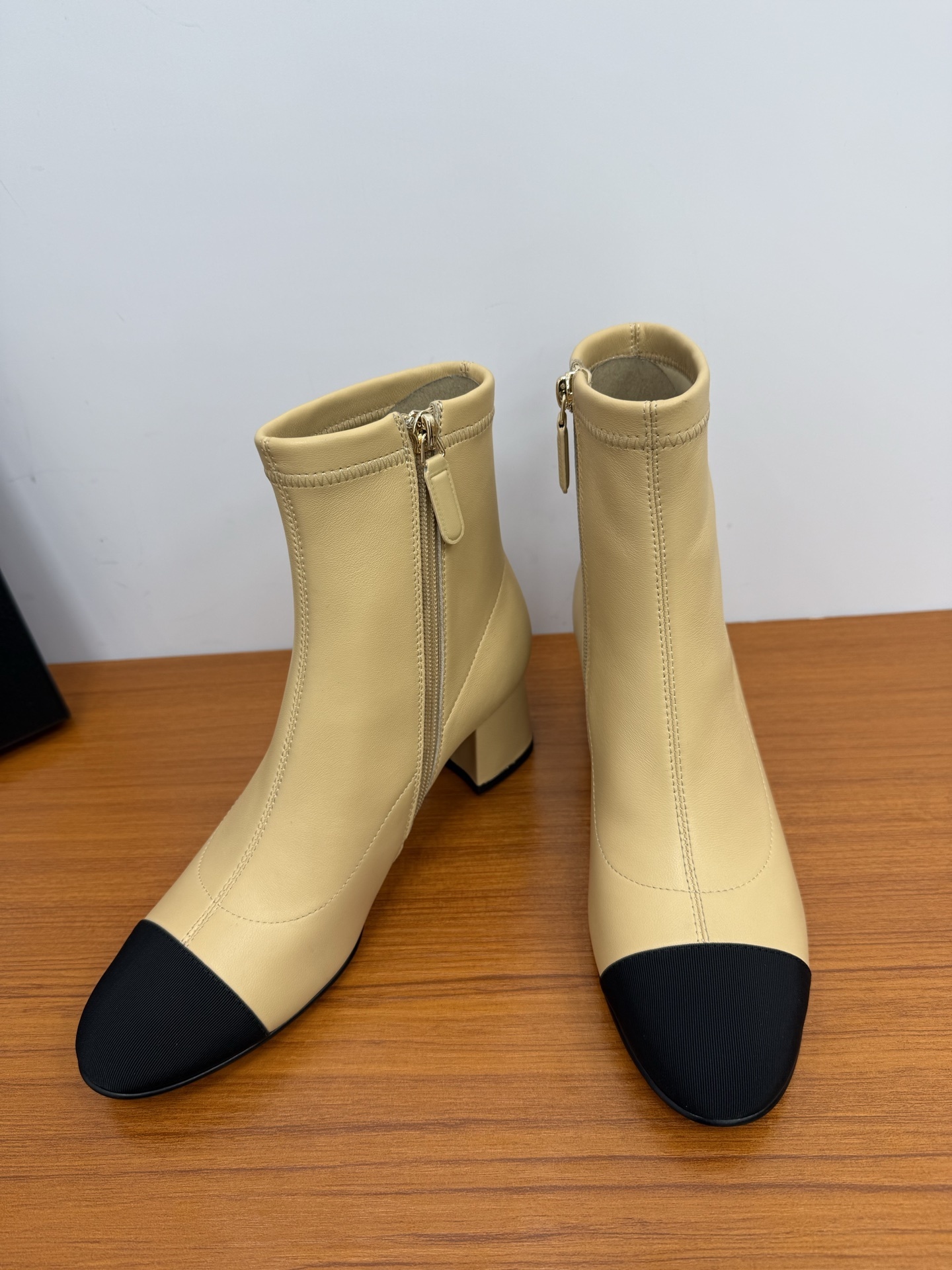 A pair of elegant beige leather-look ankle boots with a black cap toe and a sturdy block heel on a wooden surface. - Hình 7