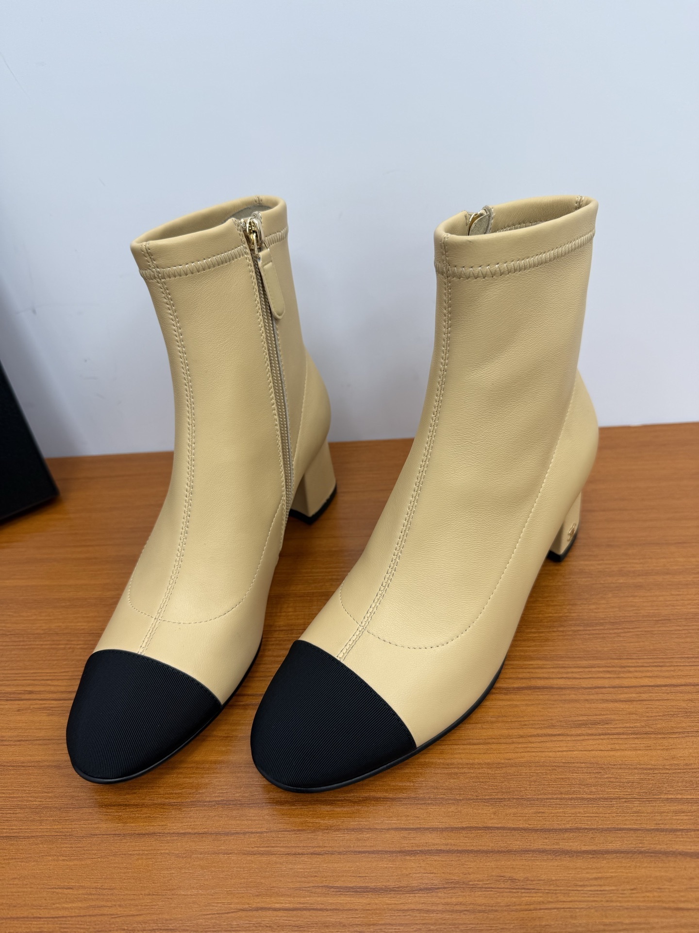 A pair of elegant beige leather-look ankle boots with a black cap toe and a sturdy block heel on a wooden surface. - Hình 5