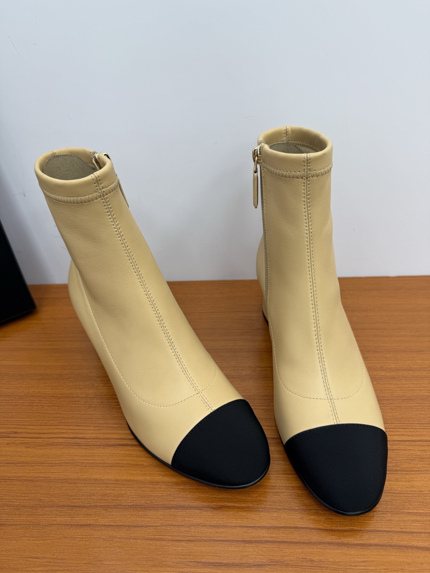 A pair of elegant beige leather-look ankle boots with a black cap toe and a sturdy block heel on a wooden surface. - Hình 4