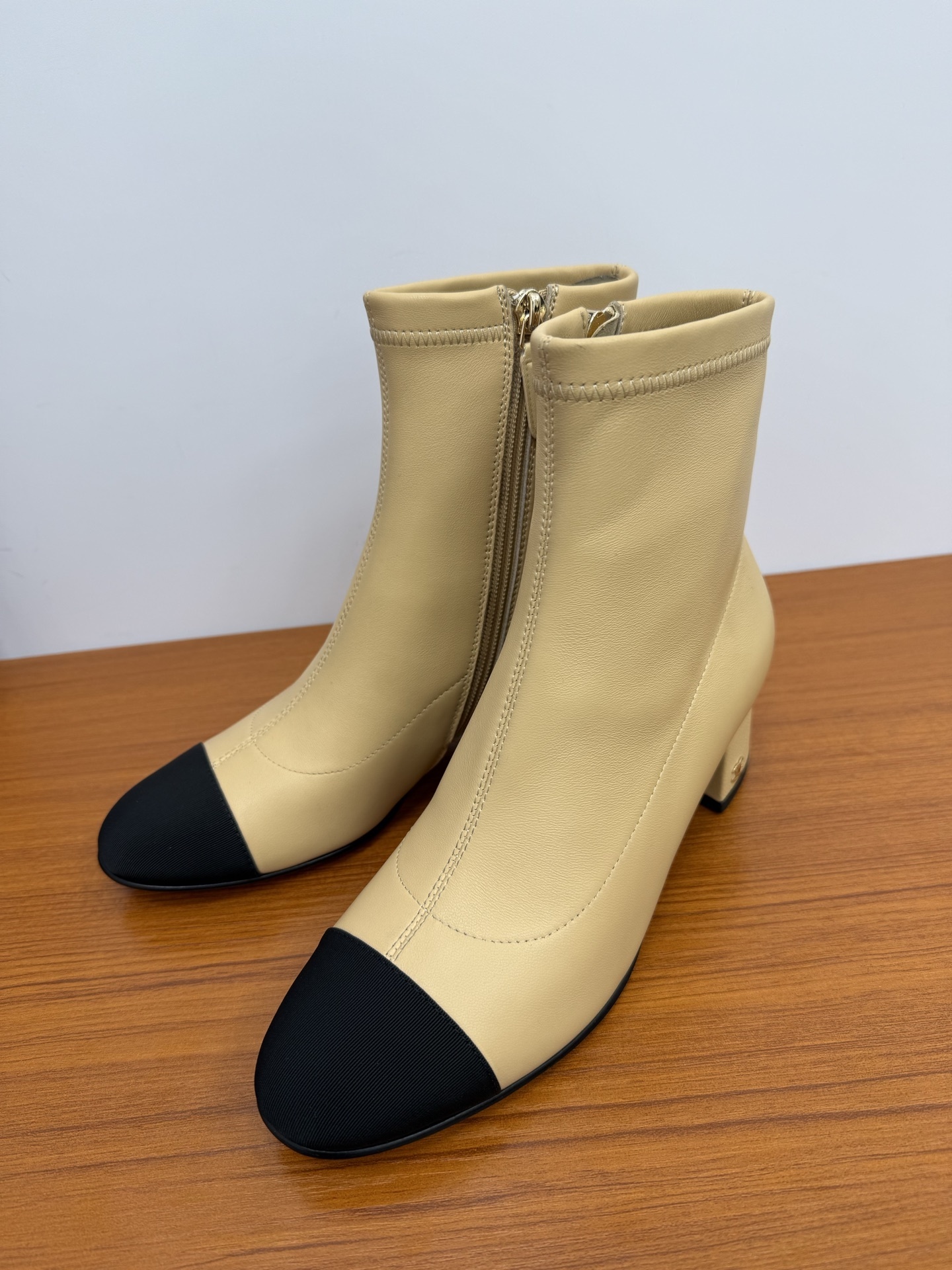 A pair of elegant beige leather-look ankle boots with a black cap toe and a sturdy block heel on a wooden surface. - Hình 3