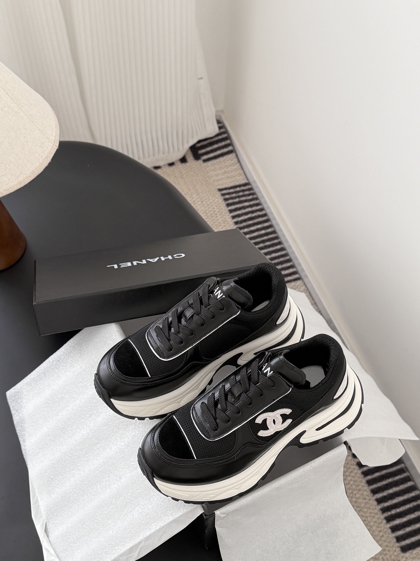 A pair of black and white Chanel chunky leather sneakers featuring the signature CC logo on a black surface next to a branded box. - Hình 9