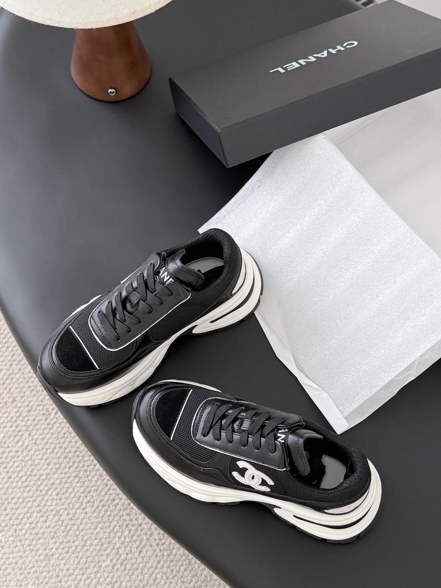 A pair of black and white Chanel chunky leather sneakers featuring the signature CC logo on a black surface next to a branded box. - Hình 6