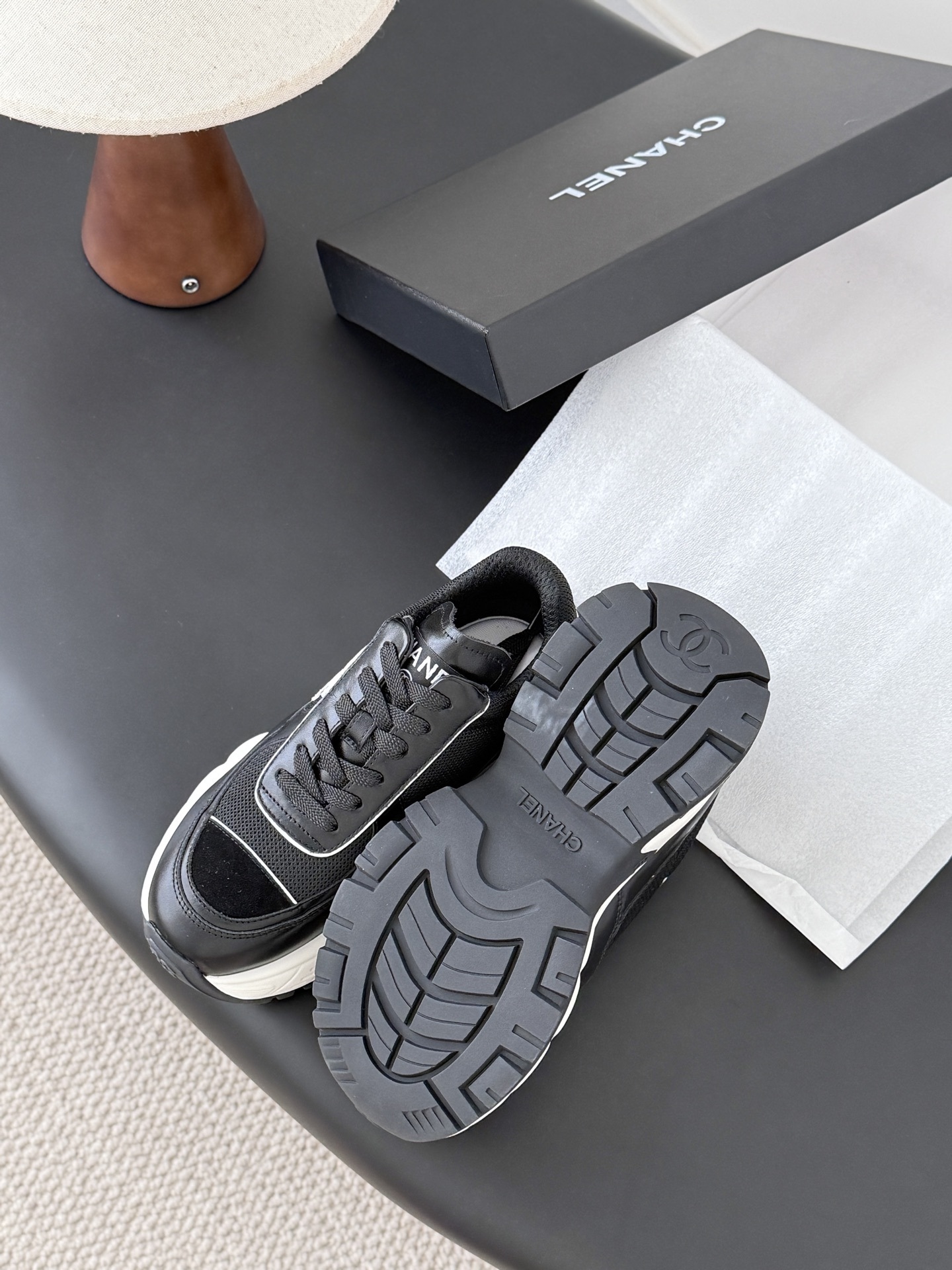 A pair of black and white Chanel chunky leather sneakers featuring the signature CC logo on a black surface next to a branded box. - Hình 5