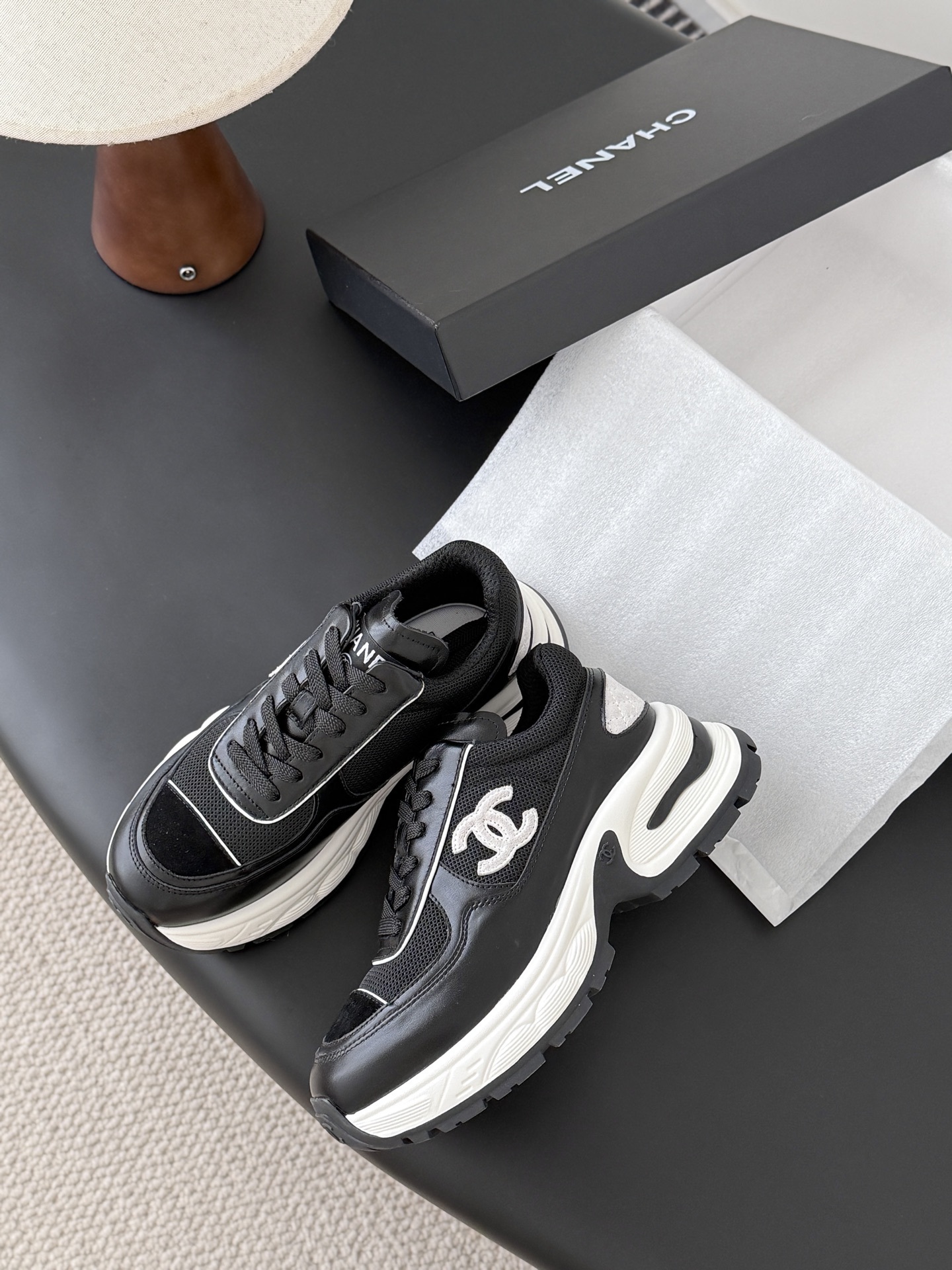 A pair of black and white Chanel chunky leather sneakers featuring the signature CC logo on a black surface next to a branded box. - Hình 4